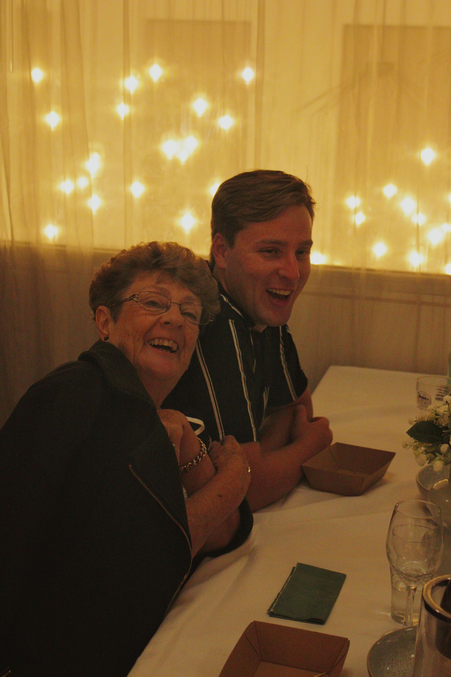 Elderly woman and young man laughing together at a decorated dinner table with fairy lights.