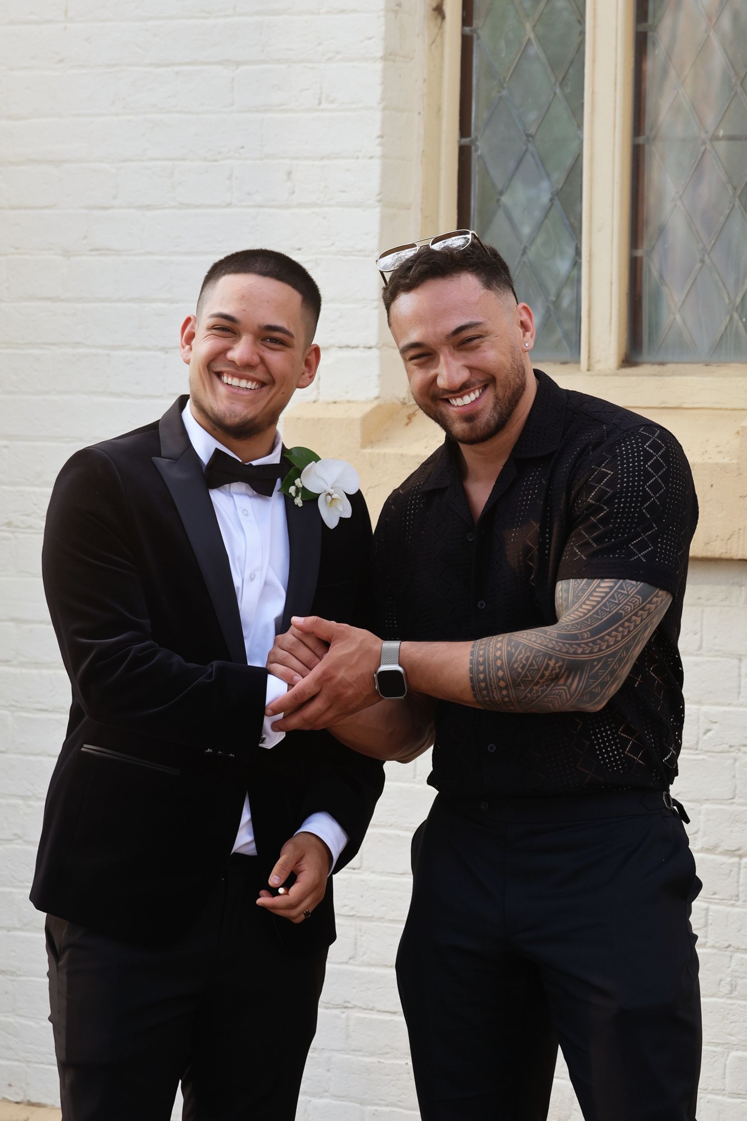 Two smiling men in formal attire pose together outside a white brick church building.