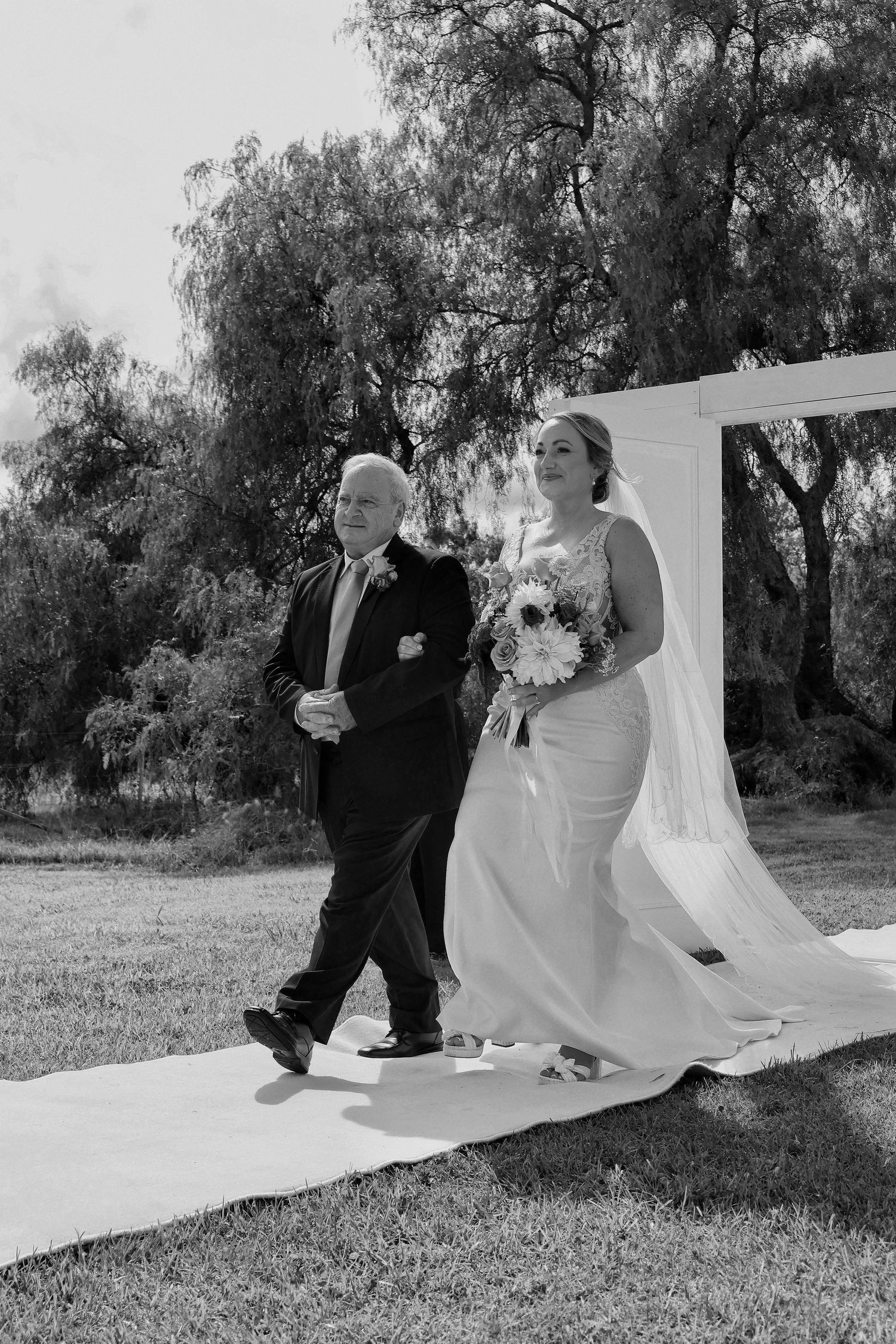 Black and white photo of bride in white gown and veil walking down aisle with older man at outdoor wedding.