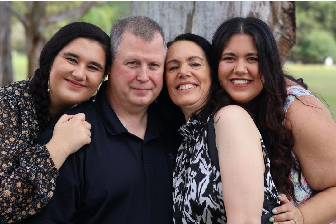 Happy family of four smiling and embracing together outdoors under shady trees.