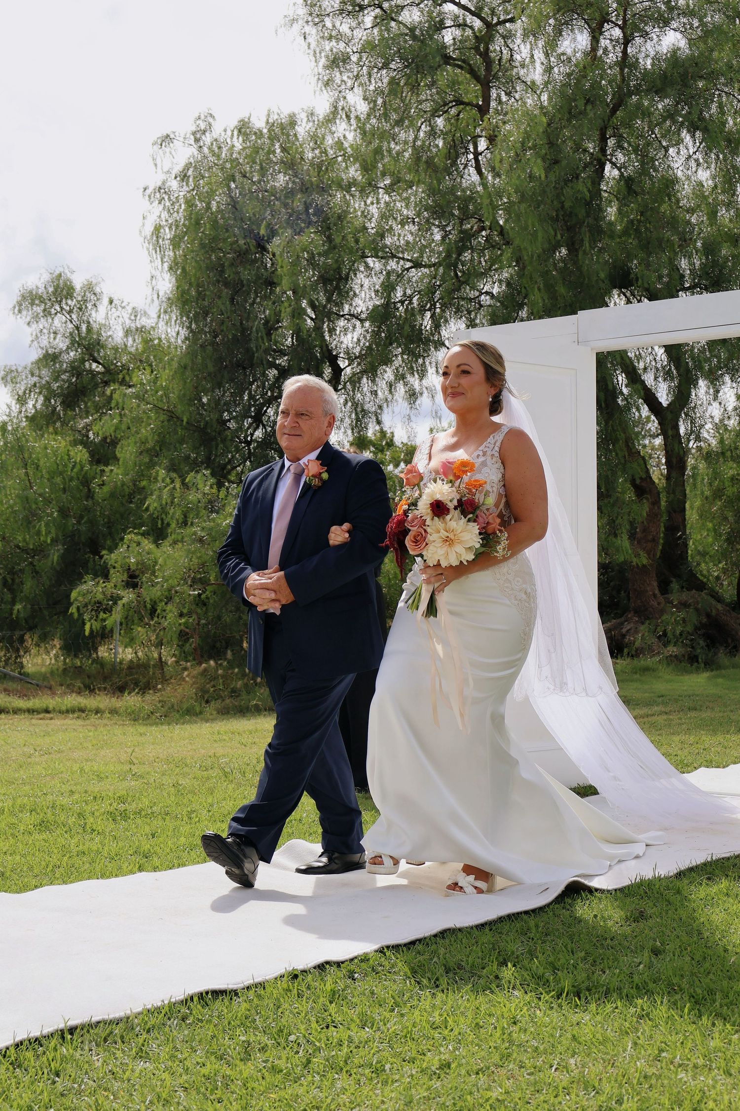 Bride walking down outdoor aisle with father, holding colorful bouquet at garden wedding.