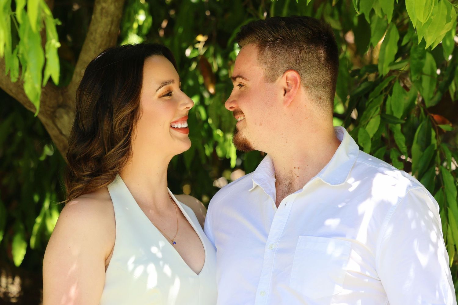 Smiling couple gazing at each other under dappled sunlight filtering through green leaves during an engagement shoot.