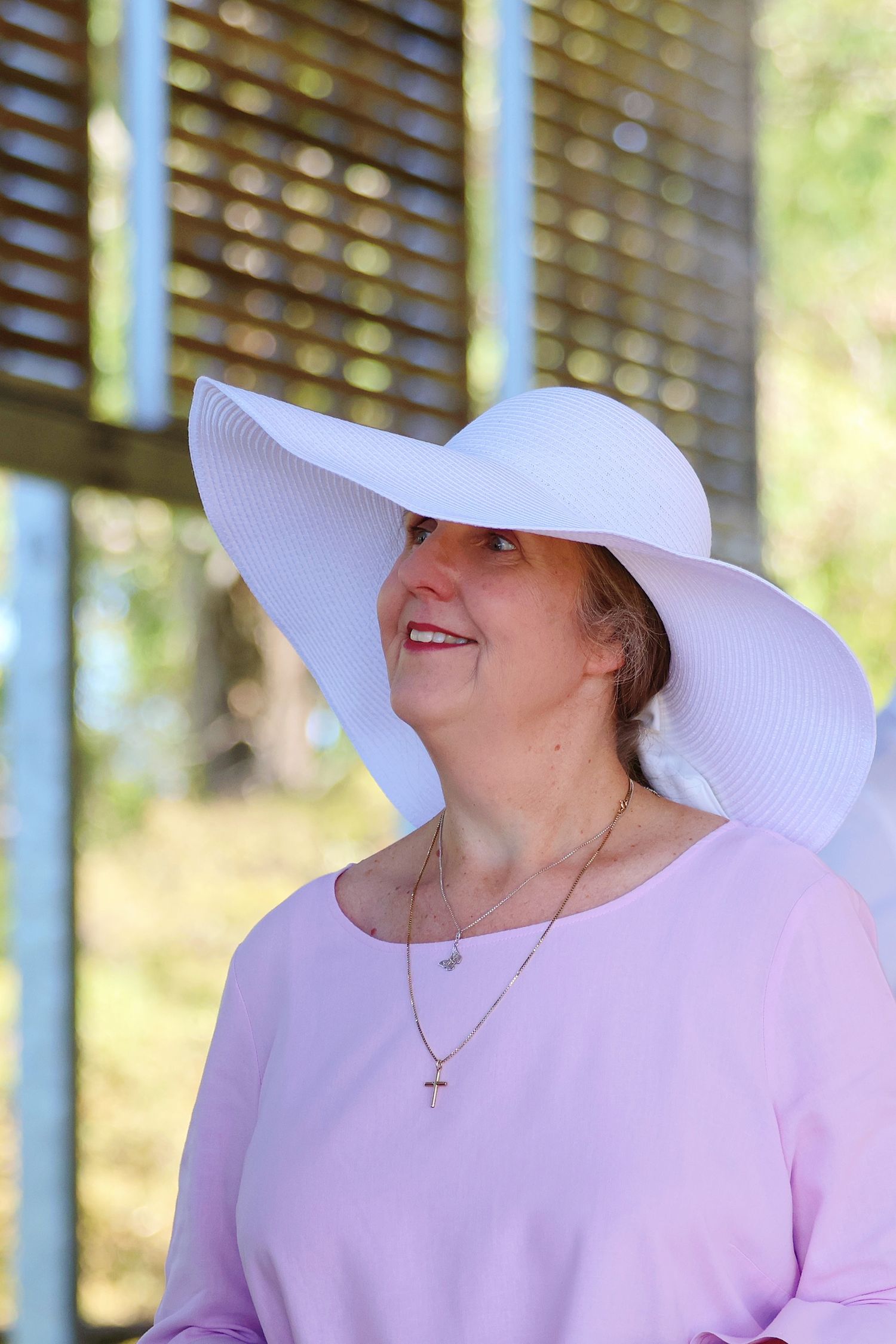Smiling middle-aged woman wearing a wide-brim white sun hat and pink top with gold cross necklace outdoors.