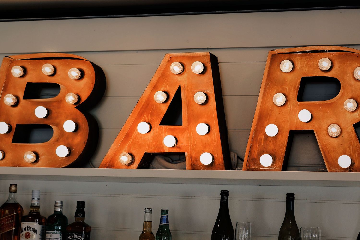 Vintage illuminated BAR marquee sign with light bulbs above shelves of liquor bottles at bar.