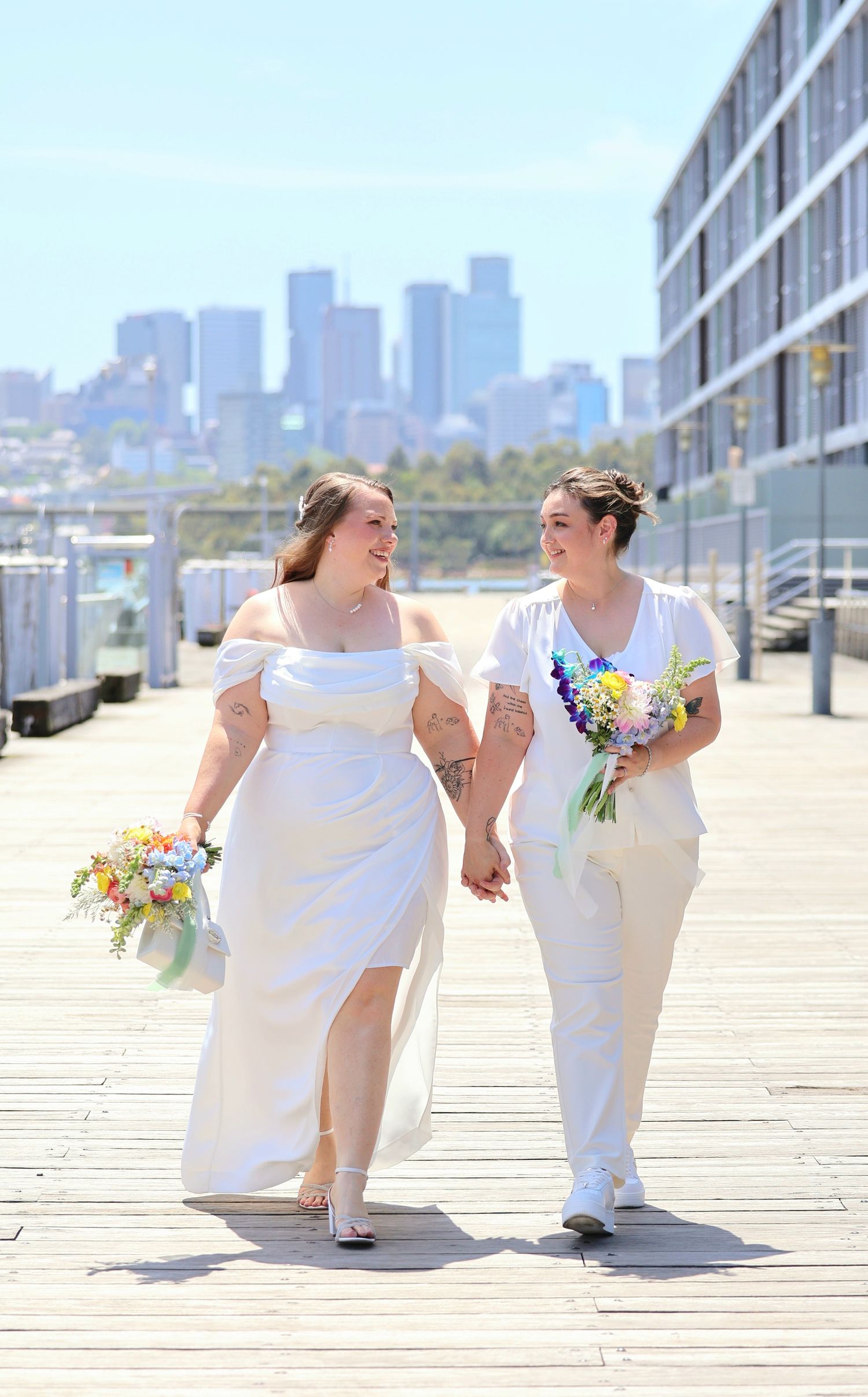 Two brides holding colorful bouquets walking hand in hand on waterfront pier with city skyline behind them.