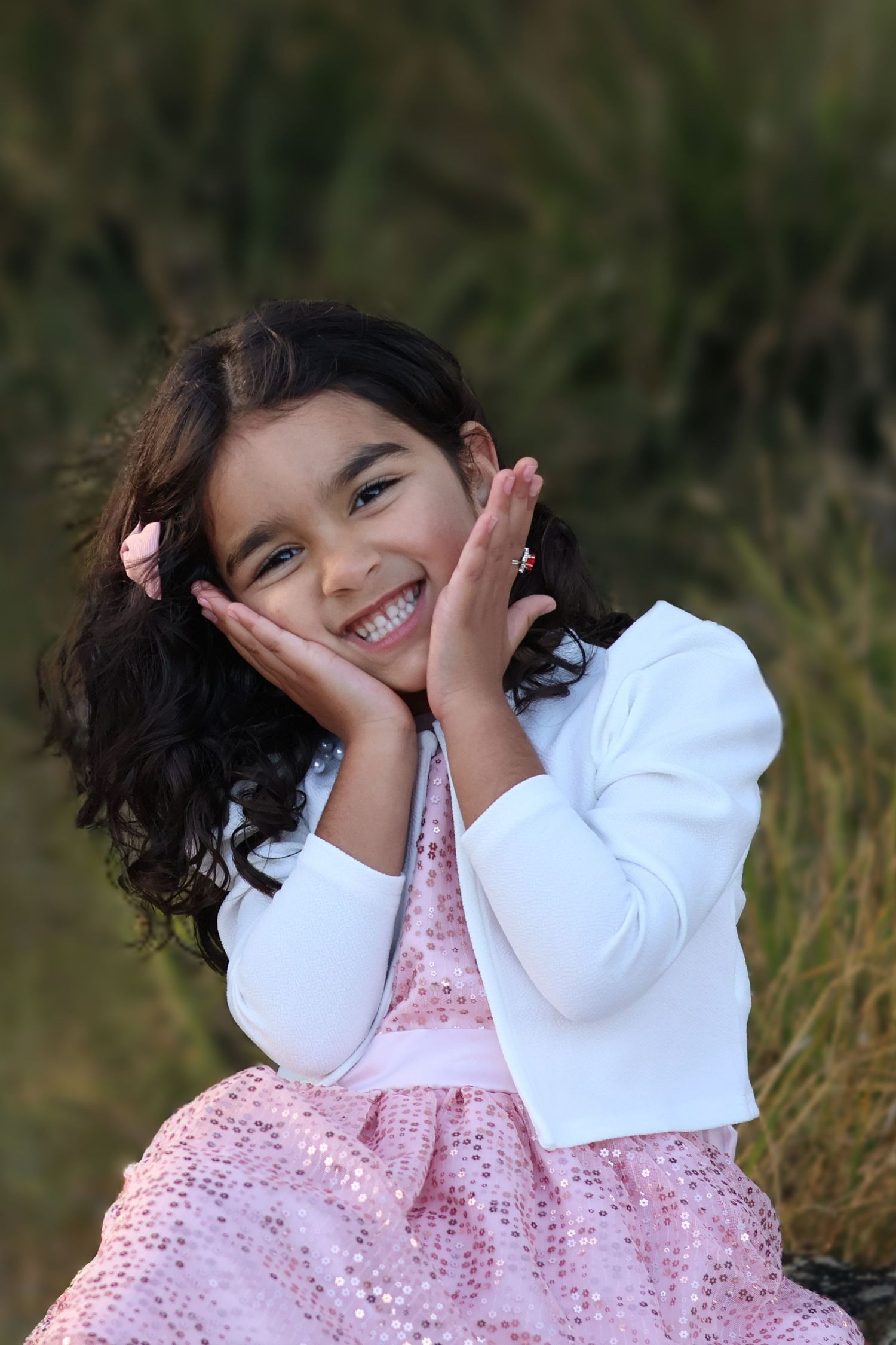 Adorable young girl in pink sequin dress and white jacket smiling with hands on cheeks.
