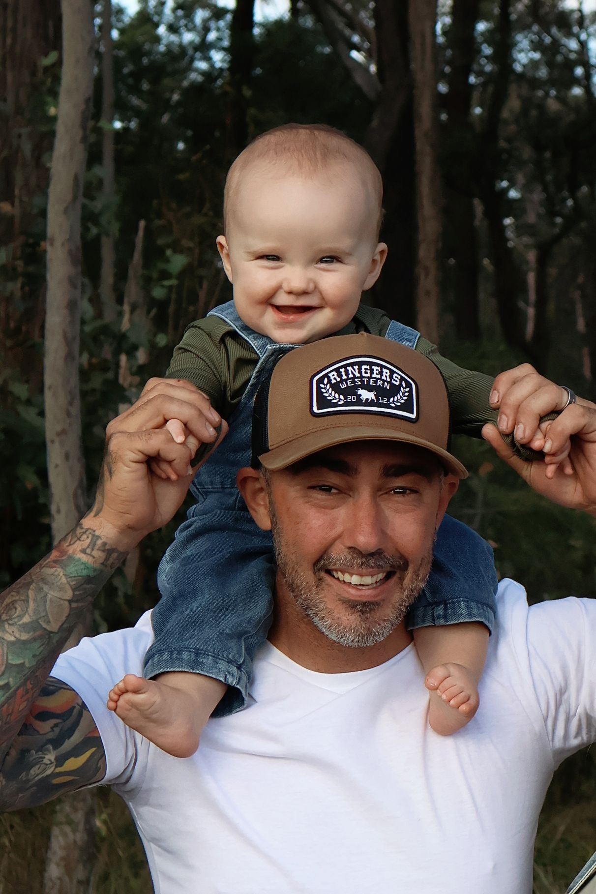Smiling baby sitting on tattooed father's shoulders outdoors in a forest, both looking happy.