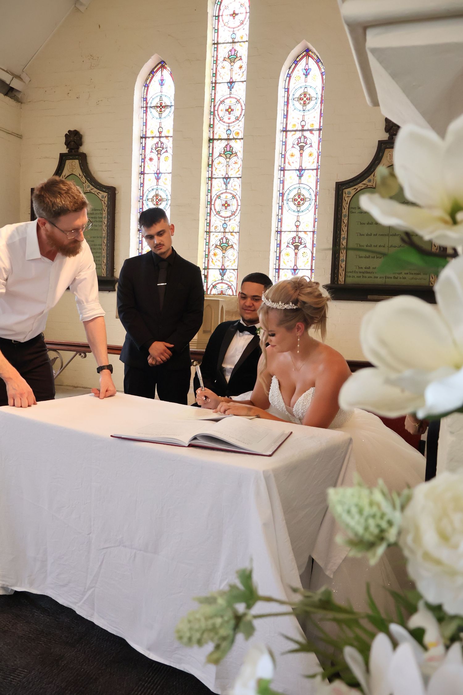 Bride signing wedding register at table while groom and witnesses look on in church.