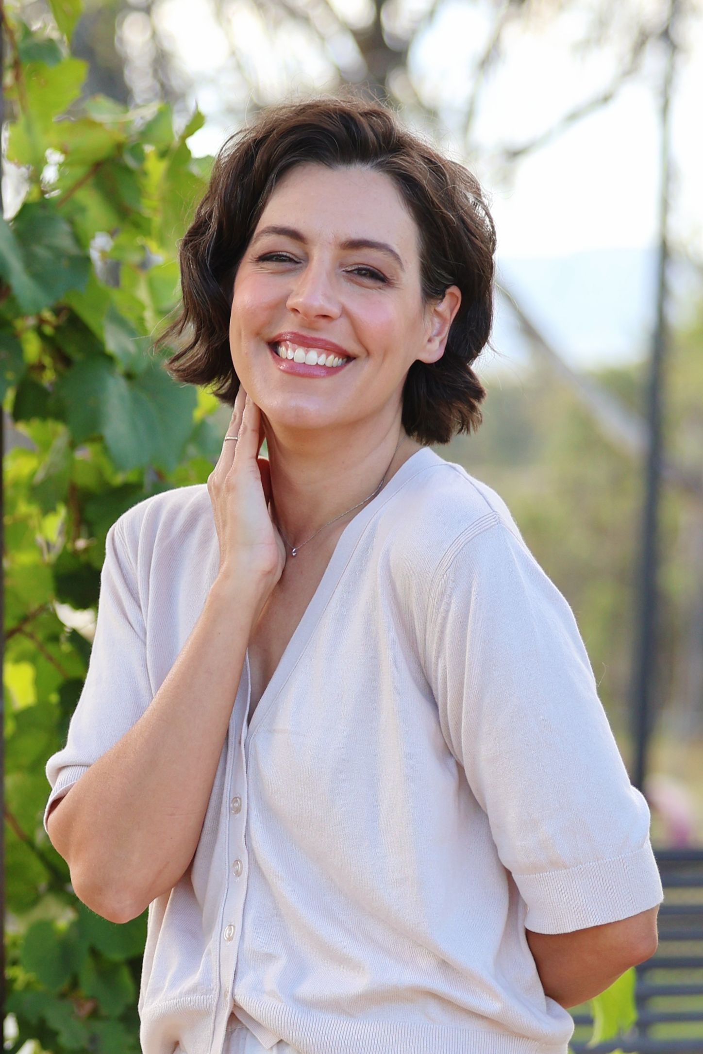 Smiling woman with short dark hair wearing a white shirt poses outdoors near green foliage.