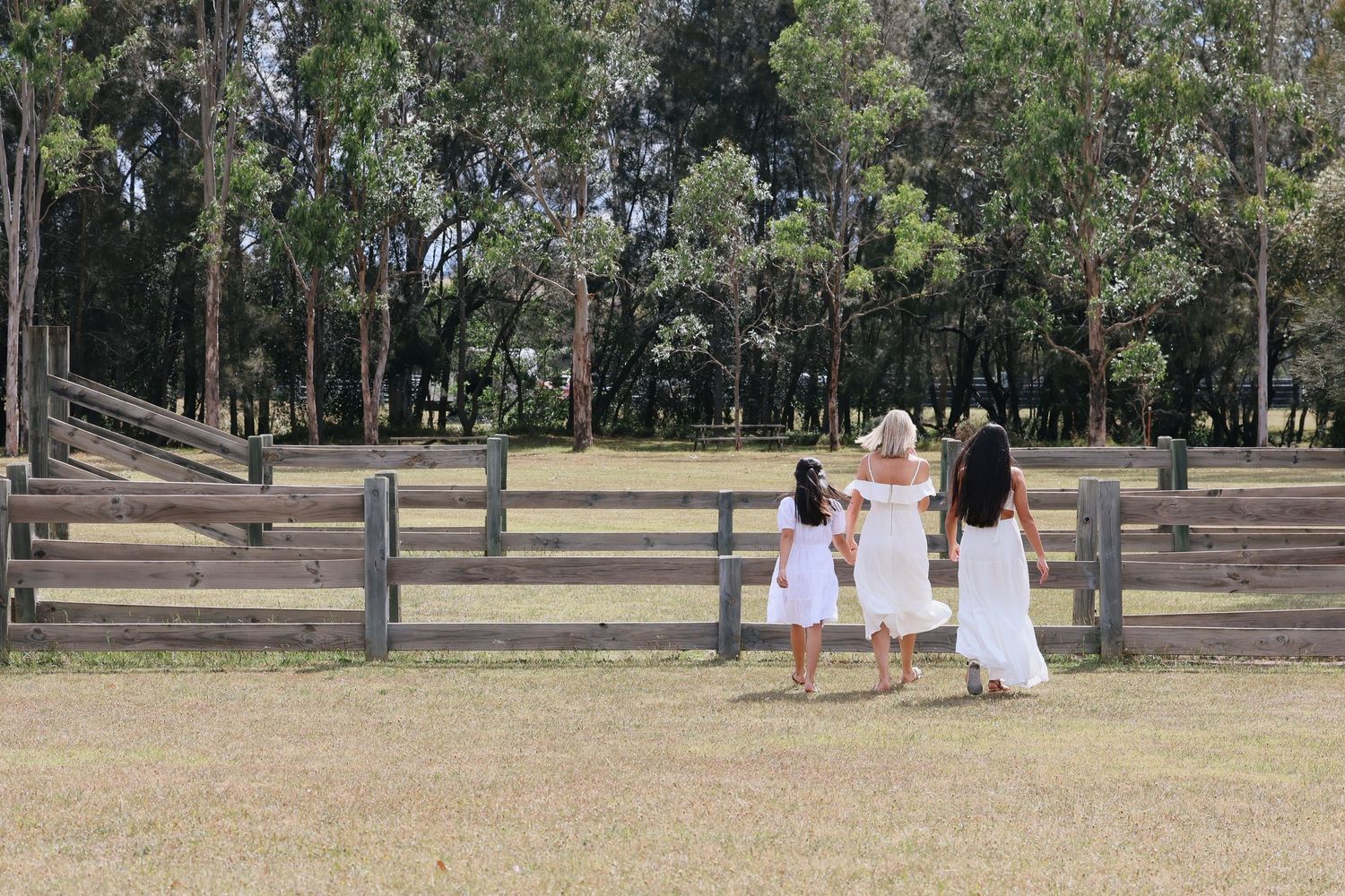 Three women in white dresses walk hand-in-hand toward a wooden fence in a lush green park surrounded by trees.