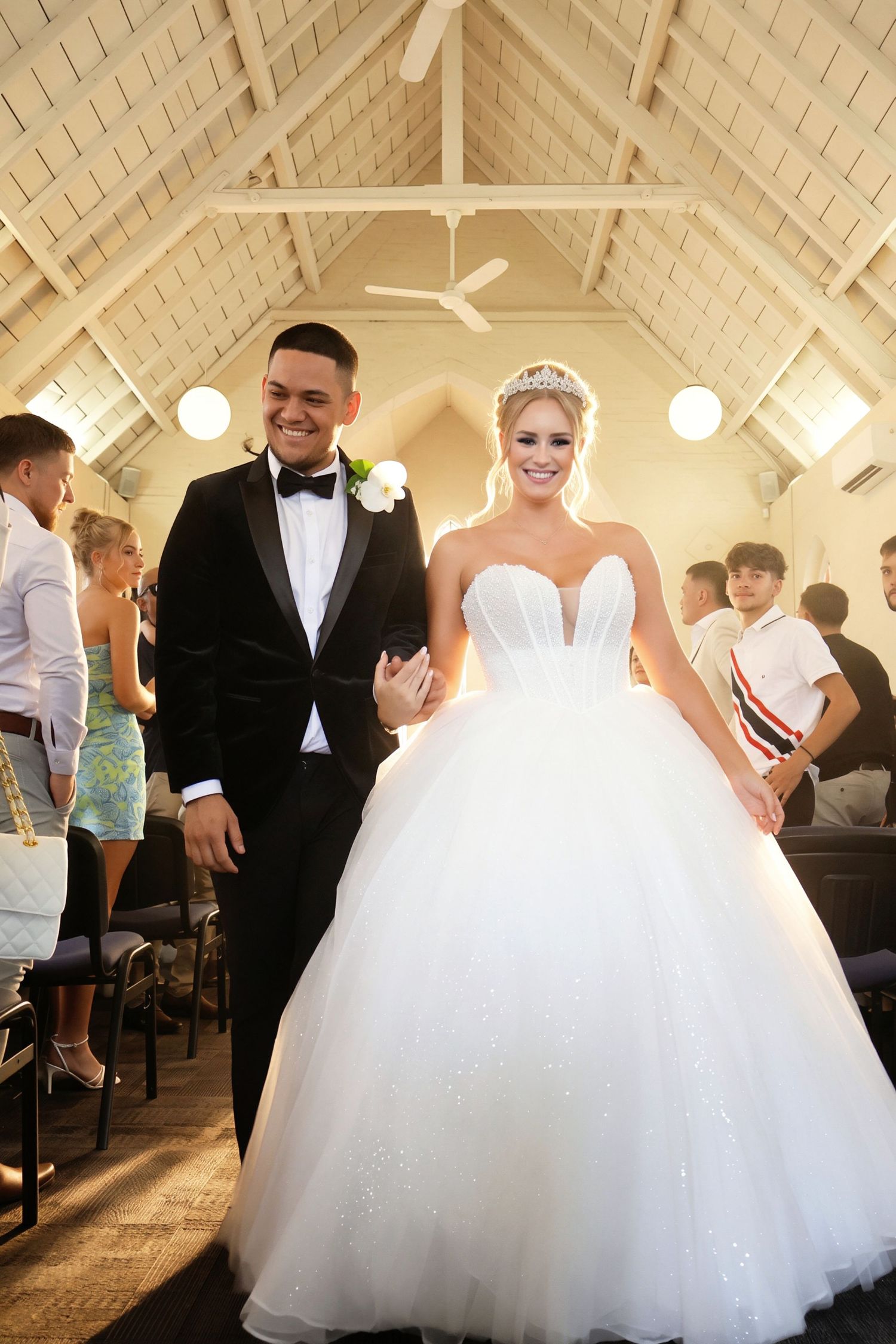Joyful newlyweds walk down church aisle, bride in sparkling ball gown with tiara, groom in tuxedo.