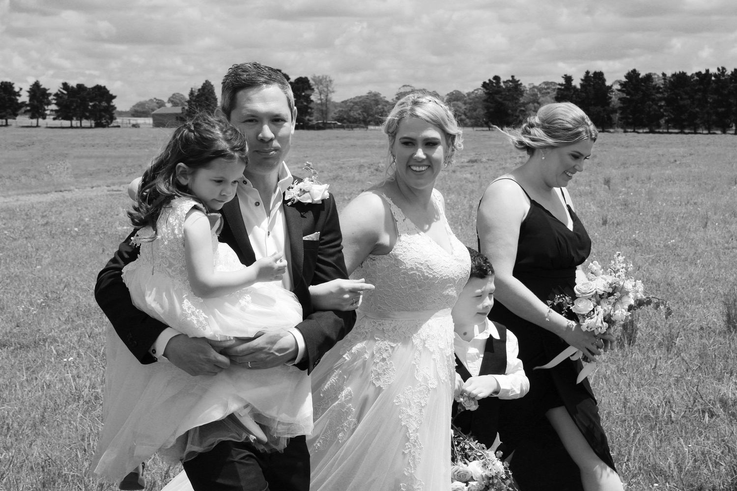 Black and white photo of groom holding flower girl with bride and bridesmaid walking across green field.