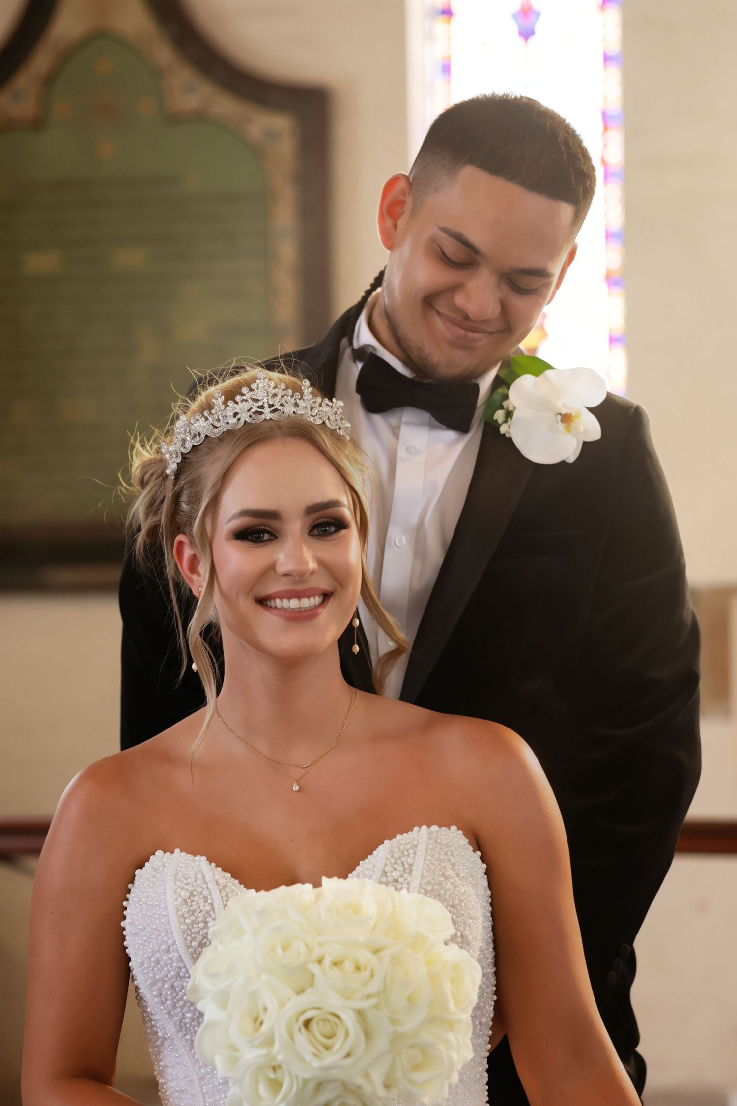 Smiling bride in beaded gown holds white rose bouquet as groom in black tuxedo stands behind her.