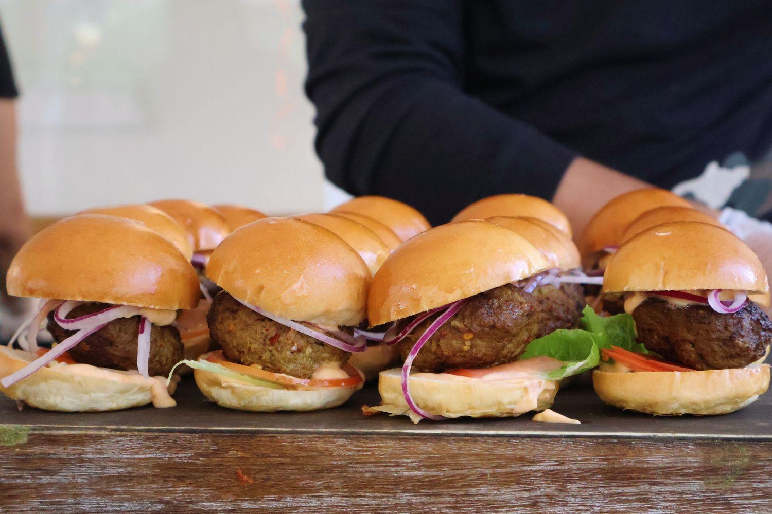 Six gourmet mini sliders with red onion and lettuce served on a wooden board.