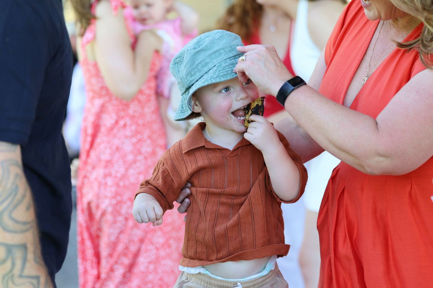 Adorable toddler in bucket hat and rust outfit being fed a treat by a woman in an orange dress.