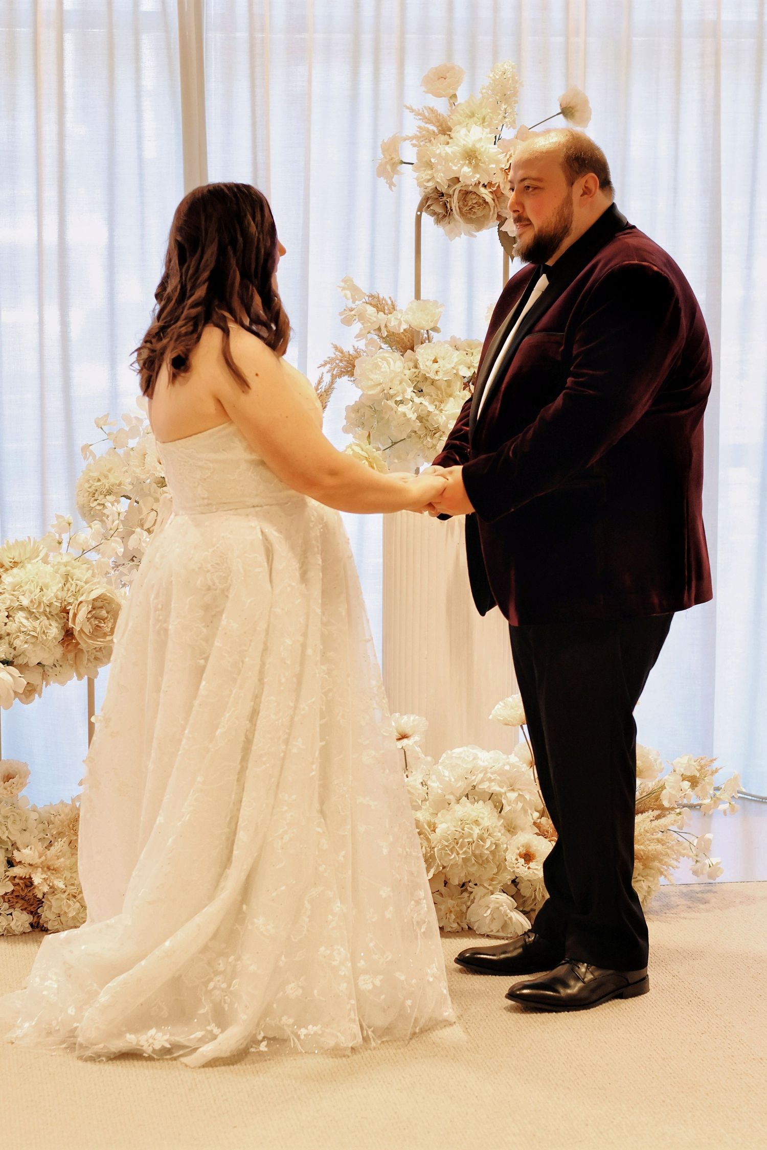 Bride and groom holding hands at altar surrounded by elegant white floral arrangements during wedding ceremony.