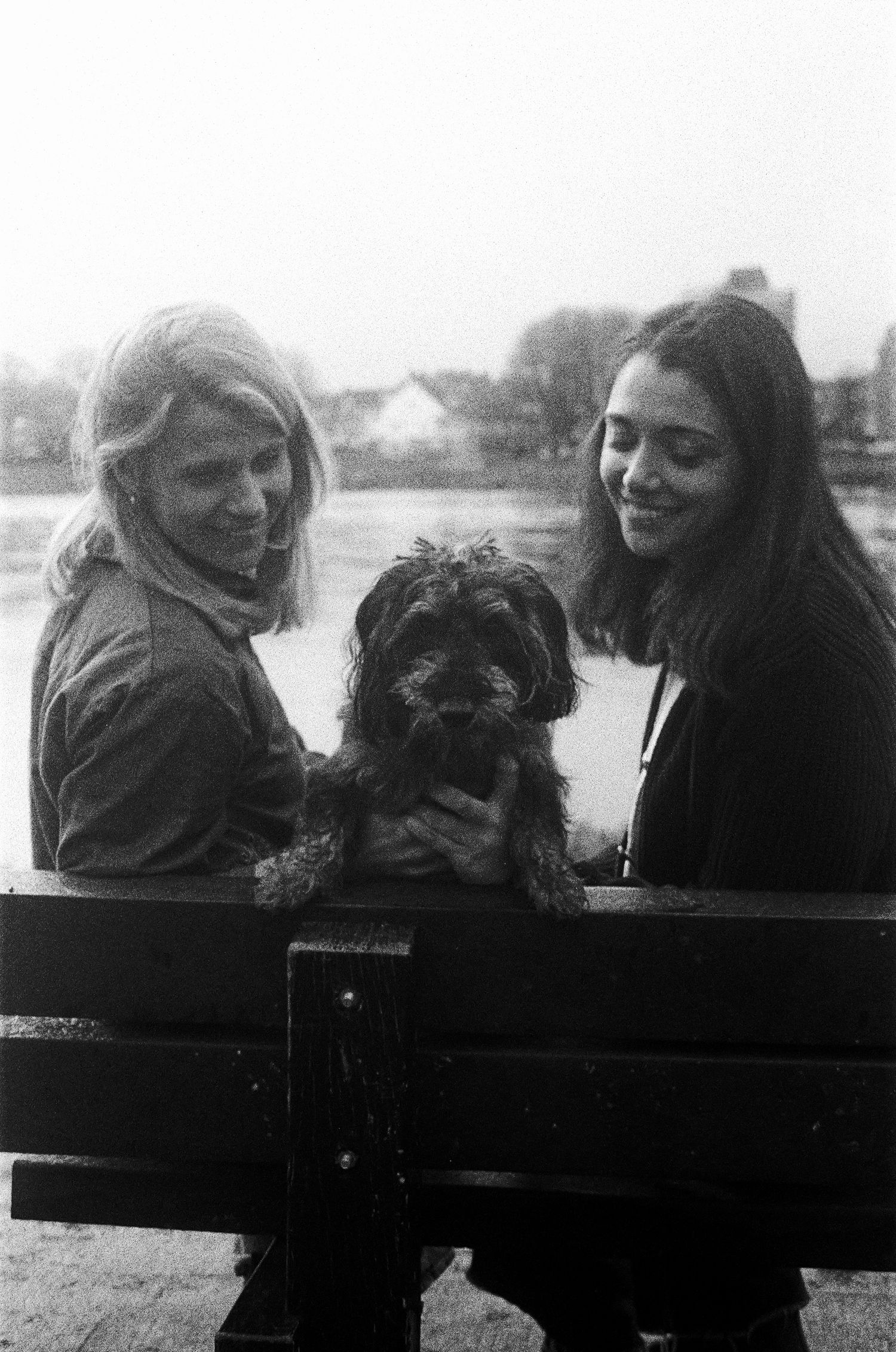 A black and white portrait 35mm film of two women with their small dachshund mix dog sitting on a bench