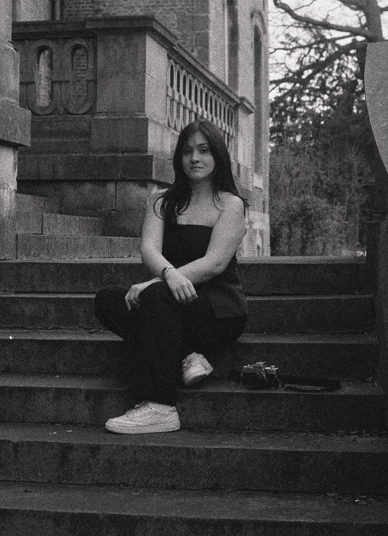 Black and white 35mm film portrait of a woman sitting on stairs with a camera next to her