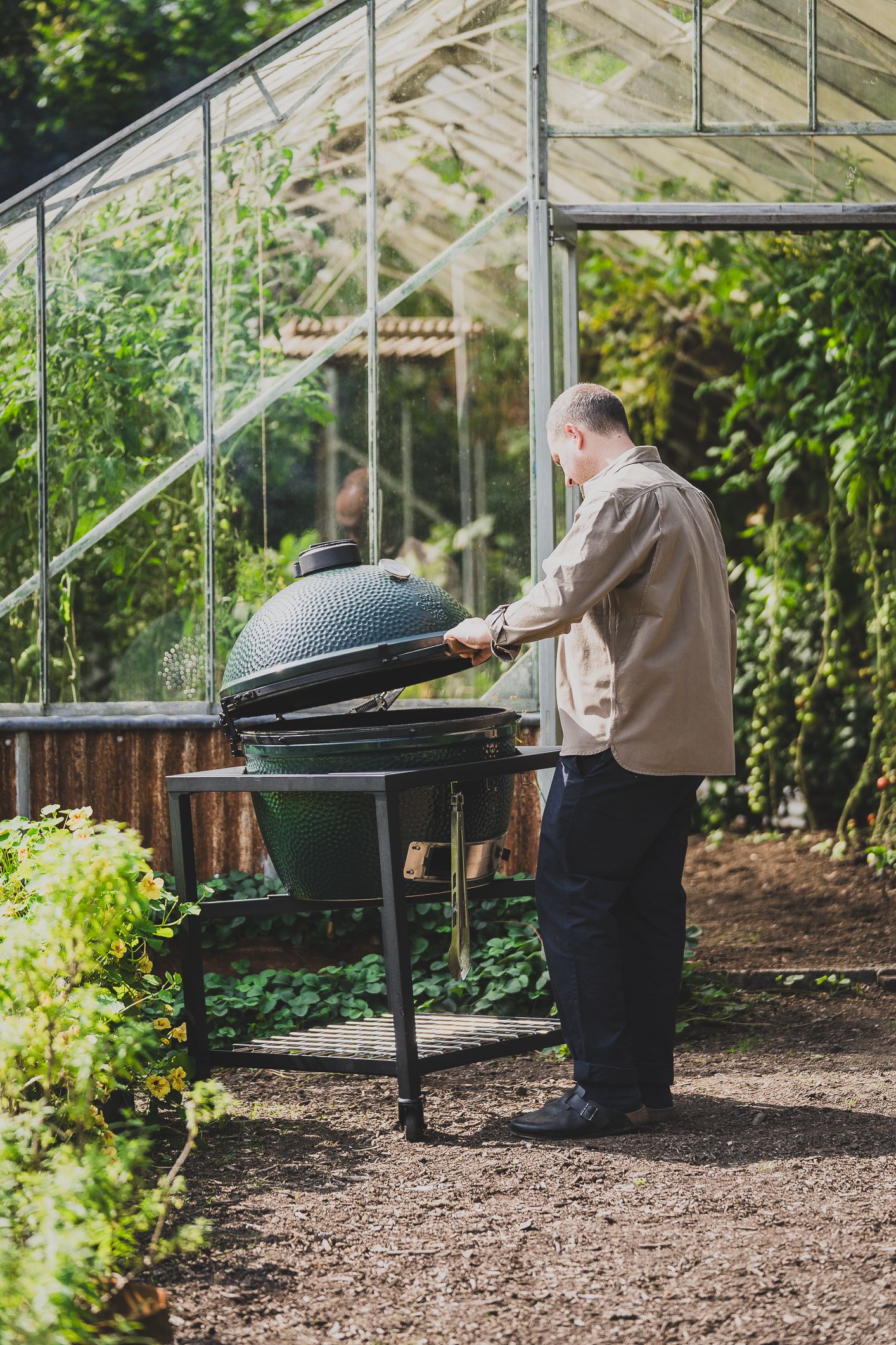 A man tending to a green Big Green Egg grill outside a glass greenhouse surrounded by lush plants.