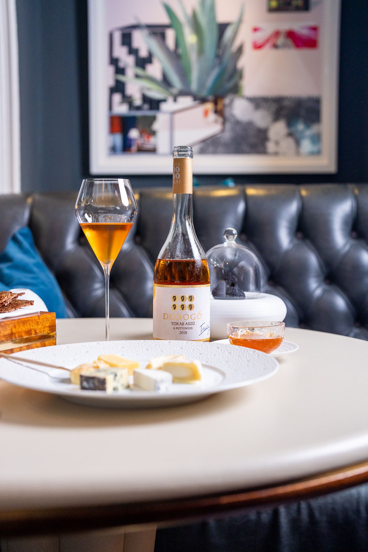 Glass of amber wine beside cheese plate and bottle on white table in stylish restaurant booth.