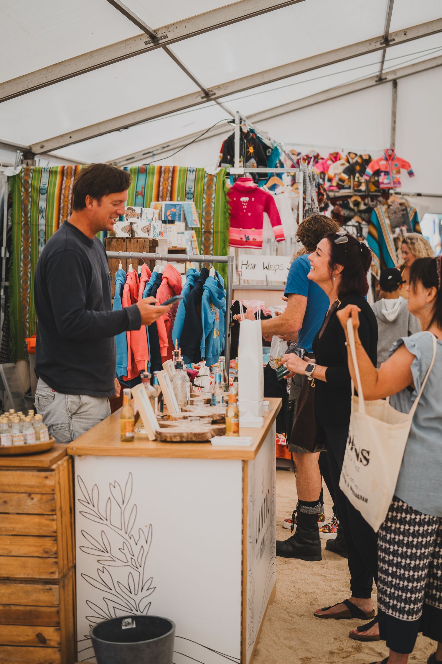 Smiling vendor assists customers at an indoor market stall with clothing and products on display.