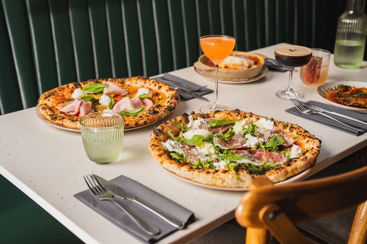 Two artisan pizzas with fresh toppings served on restaurant table with cocktails and wine.