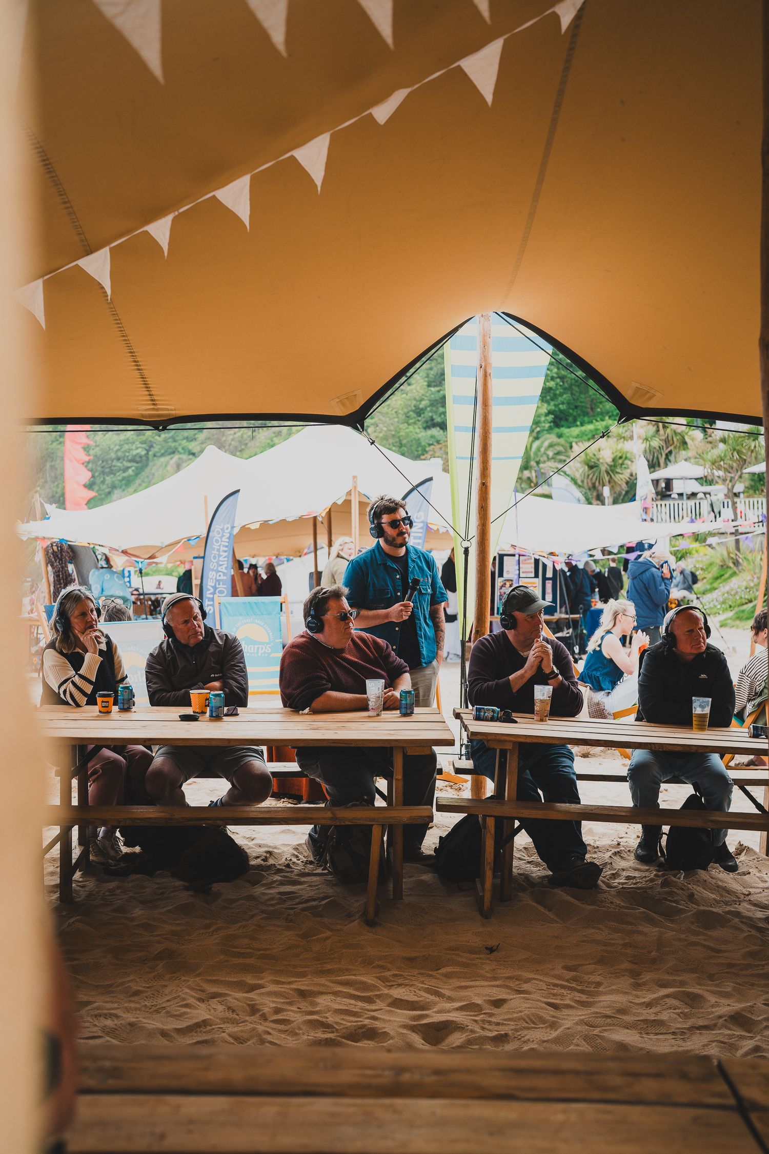 People sitting on benches inside a festival tent on sand, enjoying drinks with bunting decorations above.
