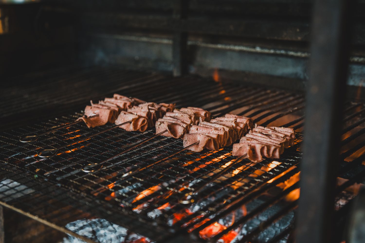 Meat skewers grilling over open flames on a charcoal barbecue grill in a smoky outdoor setting.