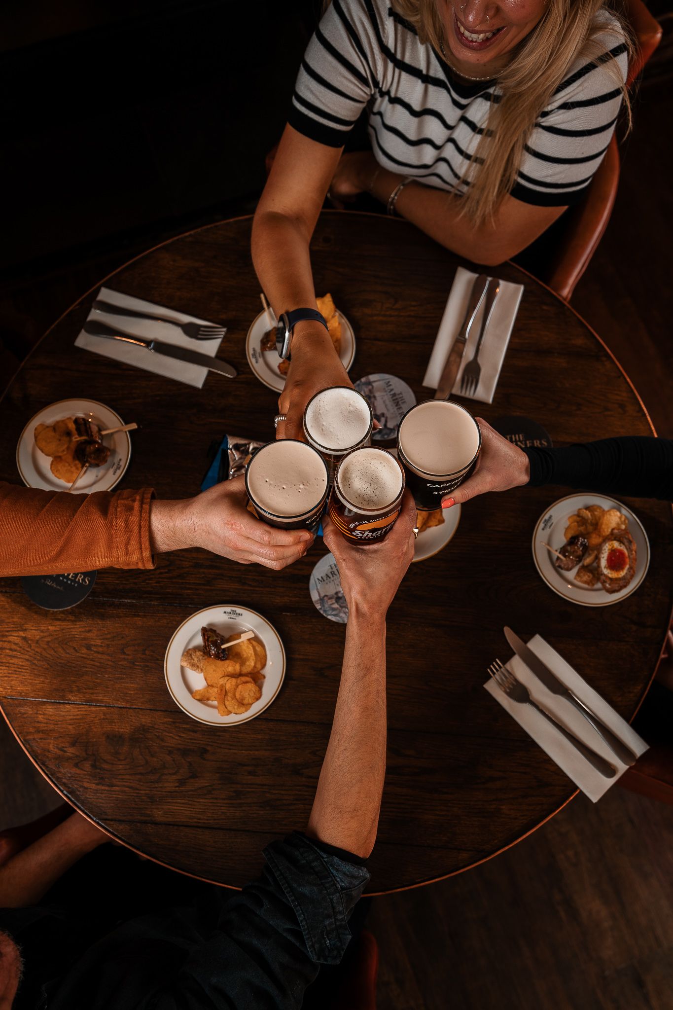 Overhead view of friends toasting with pints of beer over a round pub table with food.