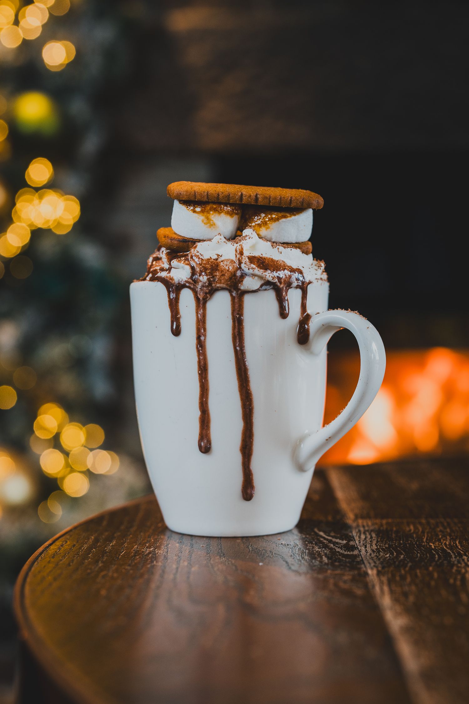 Indulgent hot chocolate topped with marshmallows and biscuit beside a glowing Christmas tree.