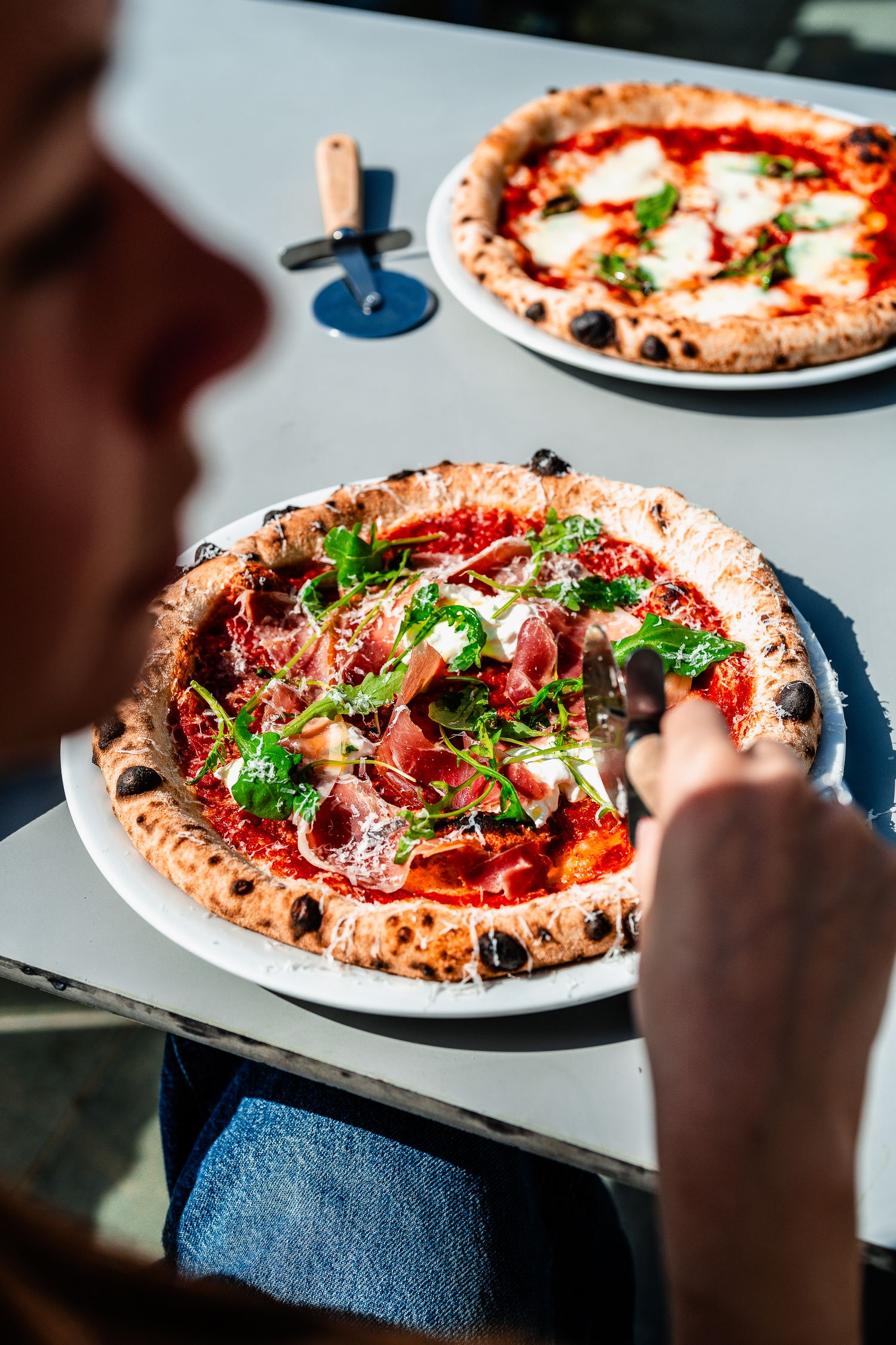 Person slicing a Neapolitan pizza topped with prosciutto, rocket, and parmesan in bright sunlight.
