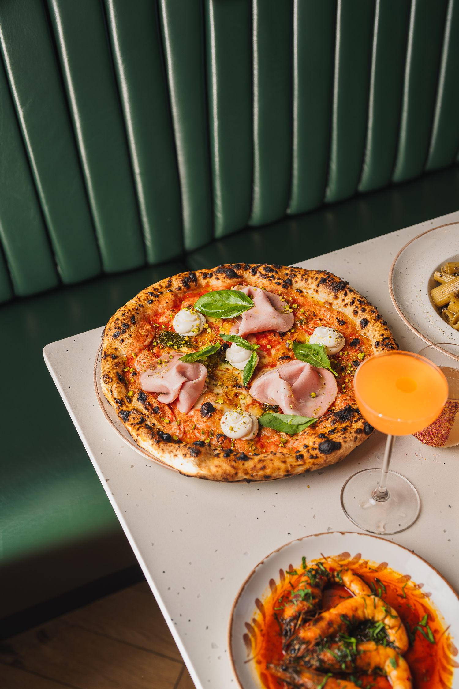 Top-down view of Neapolitan pizza with mortadella and basil next to an Aperol Spritz on a white table.