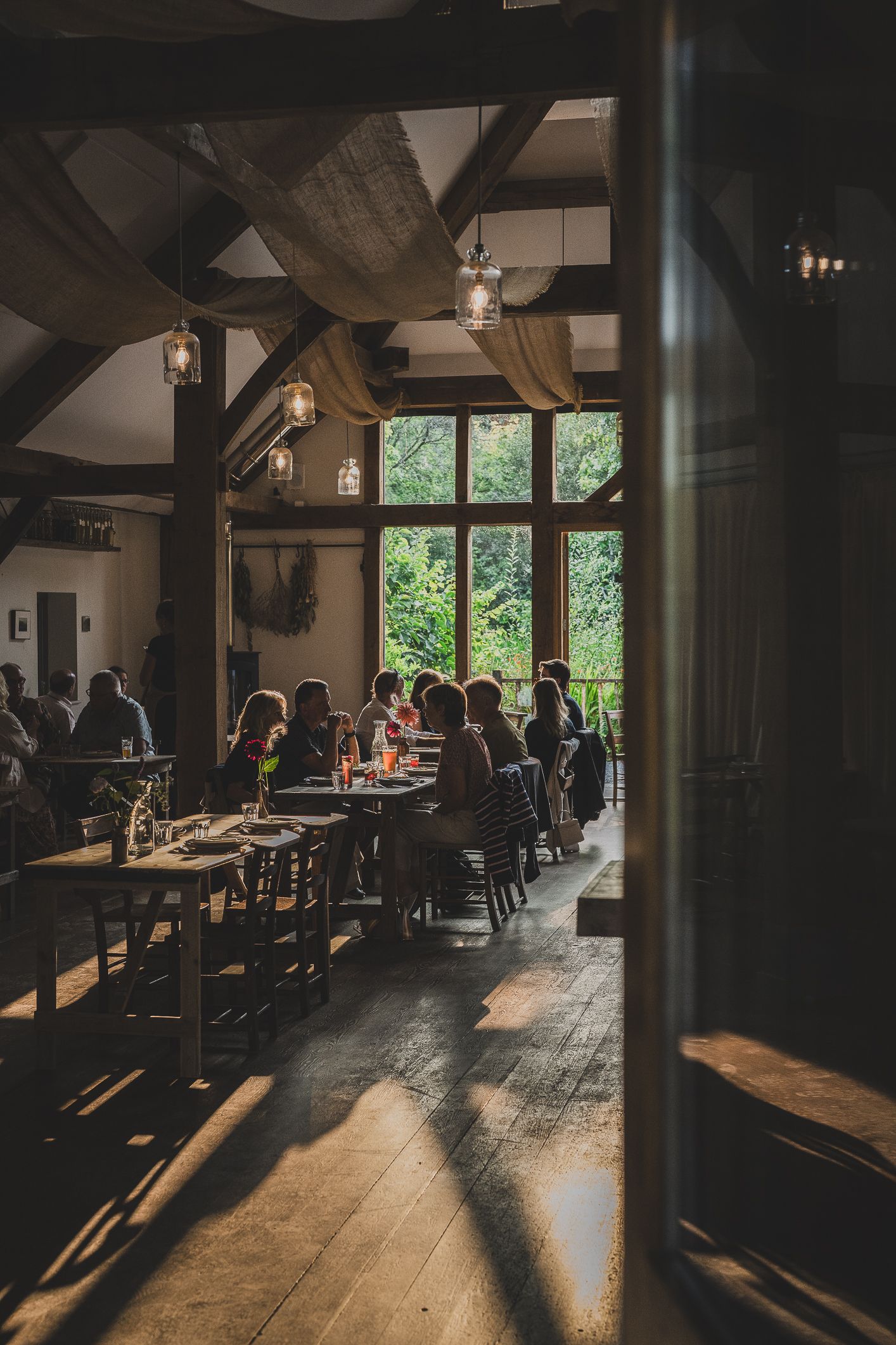 Guests dining in a warm rustic barn restaurant with wooden beams and large windows overlooking greenery.
