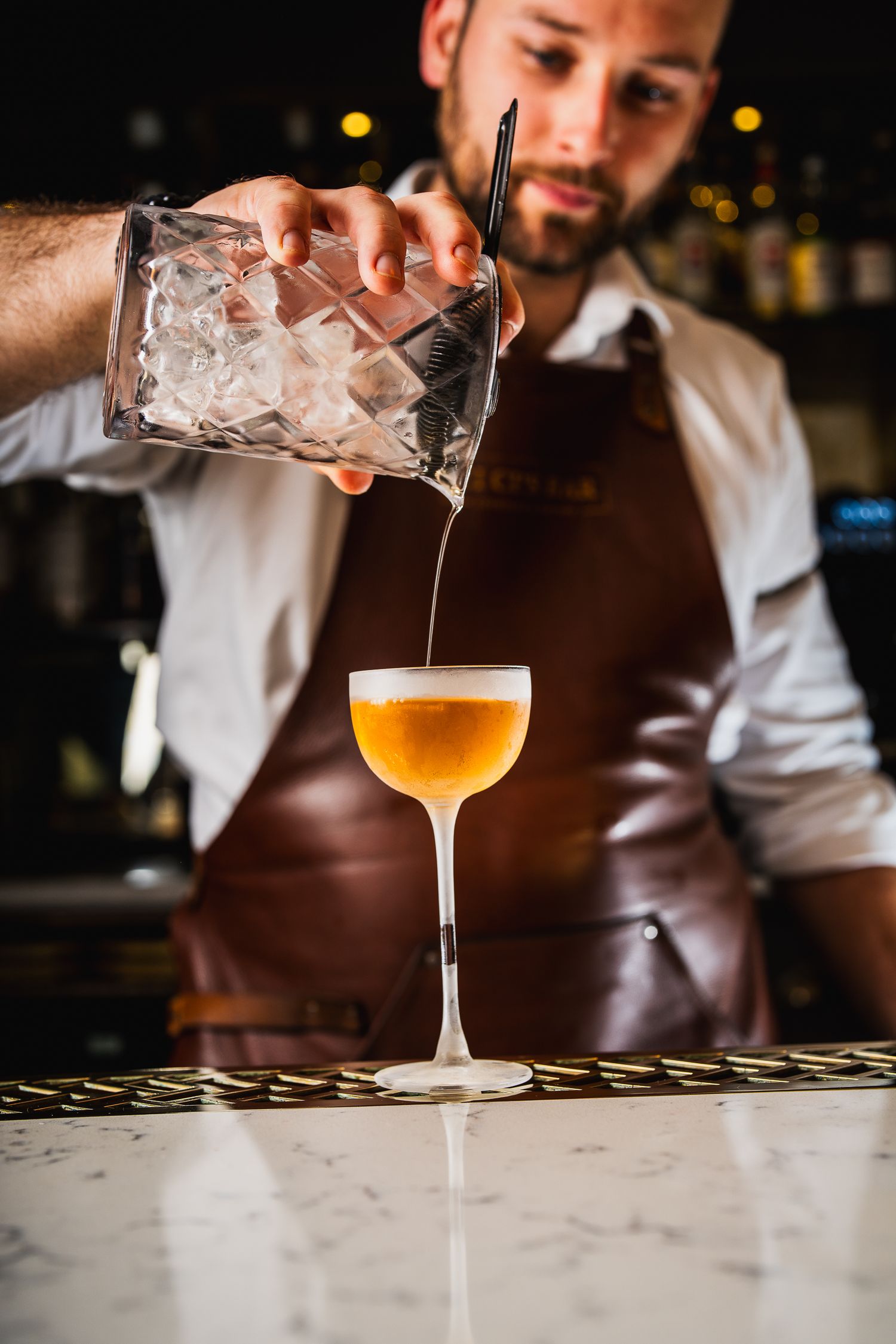 Bartender in leather apron pouring cocktail into elegant coupe glass filled with golden drink at marble bar.