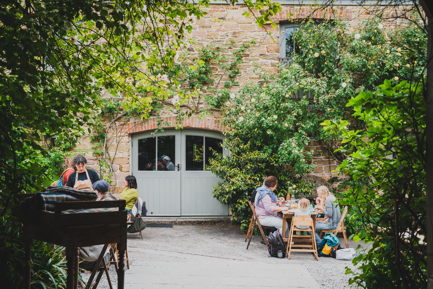 Diners enjoying an outdoor meal in a lush garden courtyard beside a charming stone building.