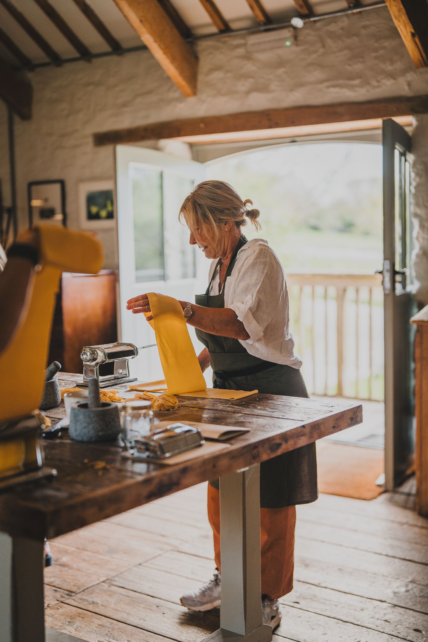 A woman in a dark apron rolls fresh yellow pasta dough using a pasta machine in a rustic kitchen.