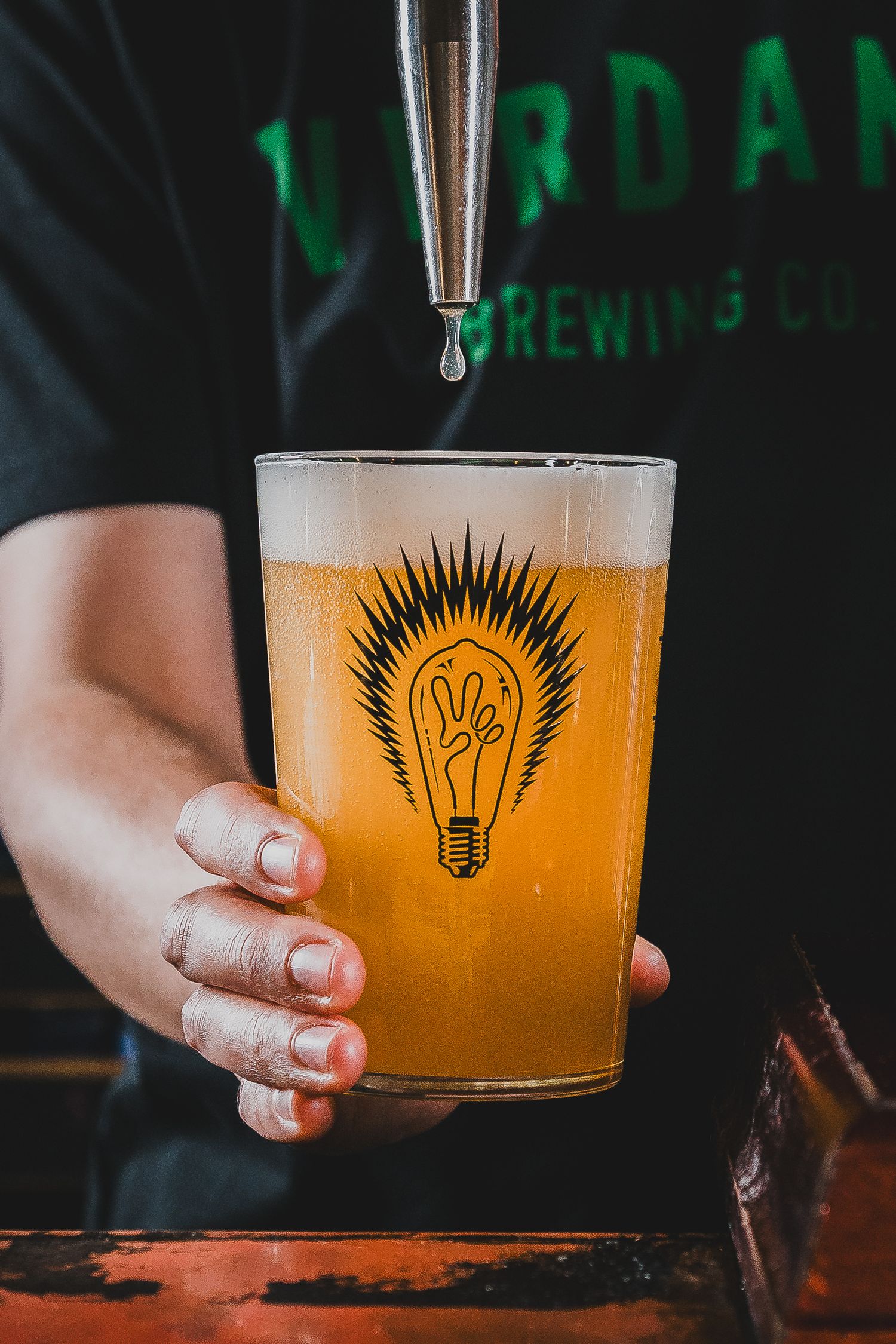 Hand holding pint glass with light bulb logo being filled with golden craft beer from tap.