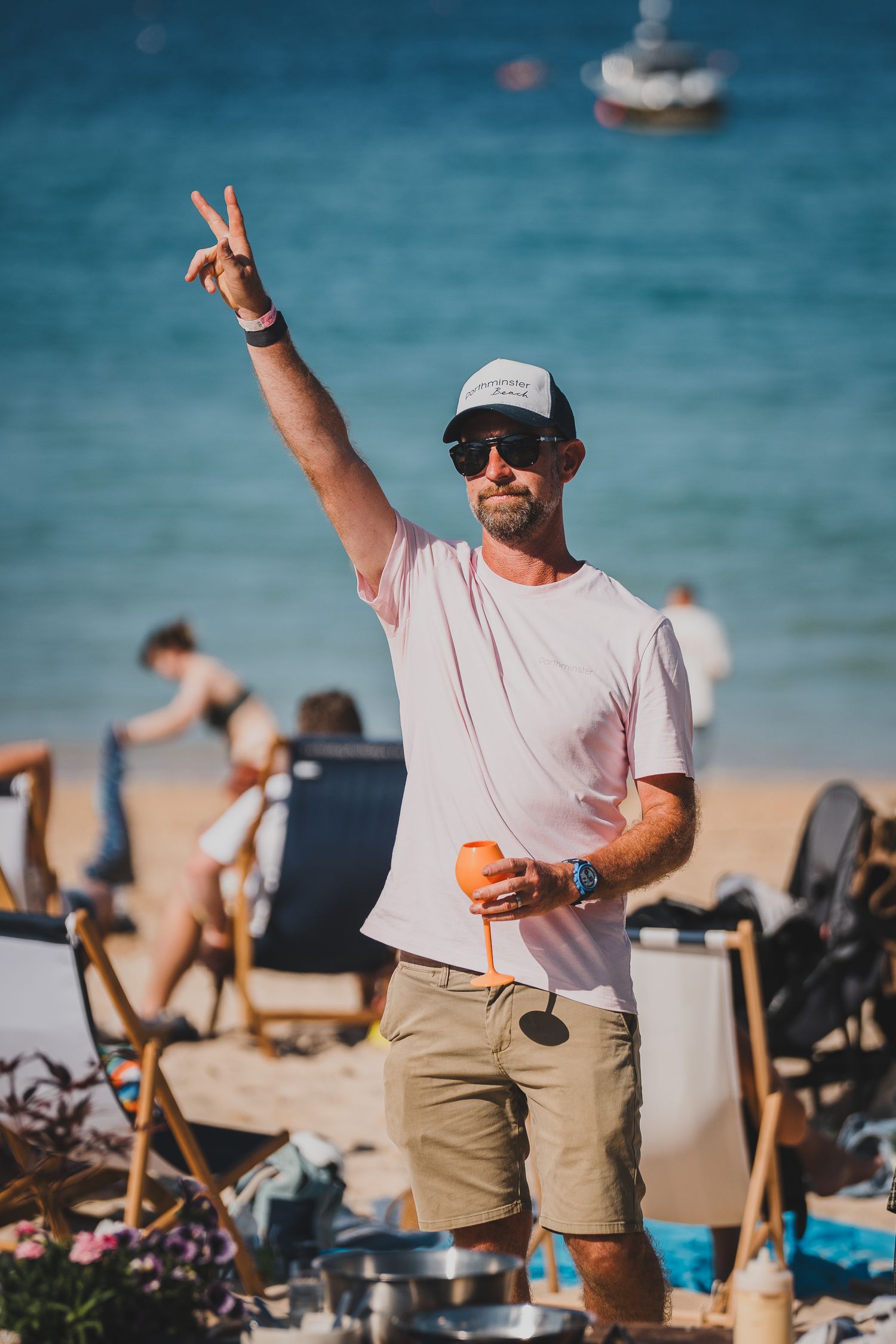 Smiling man in cap and sunglasses flashes peace sign on a sunny beach holding an orange drink.