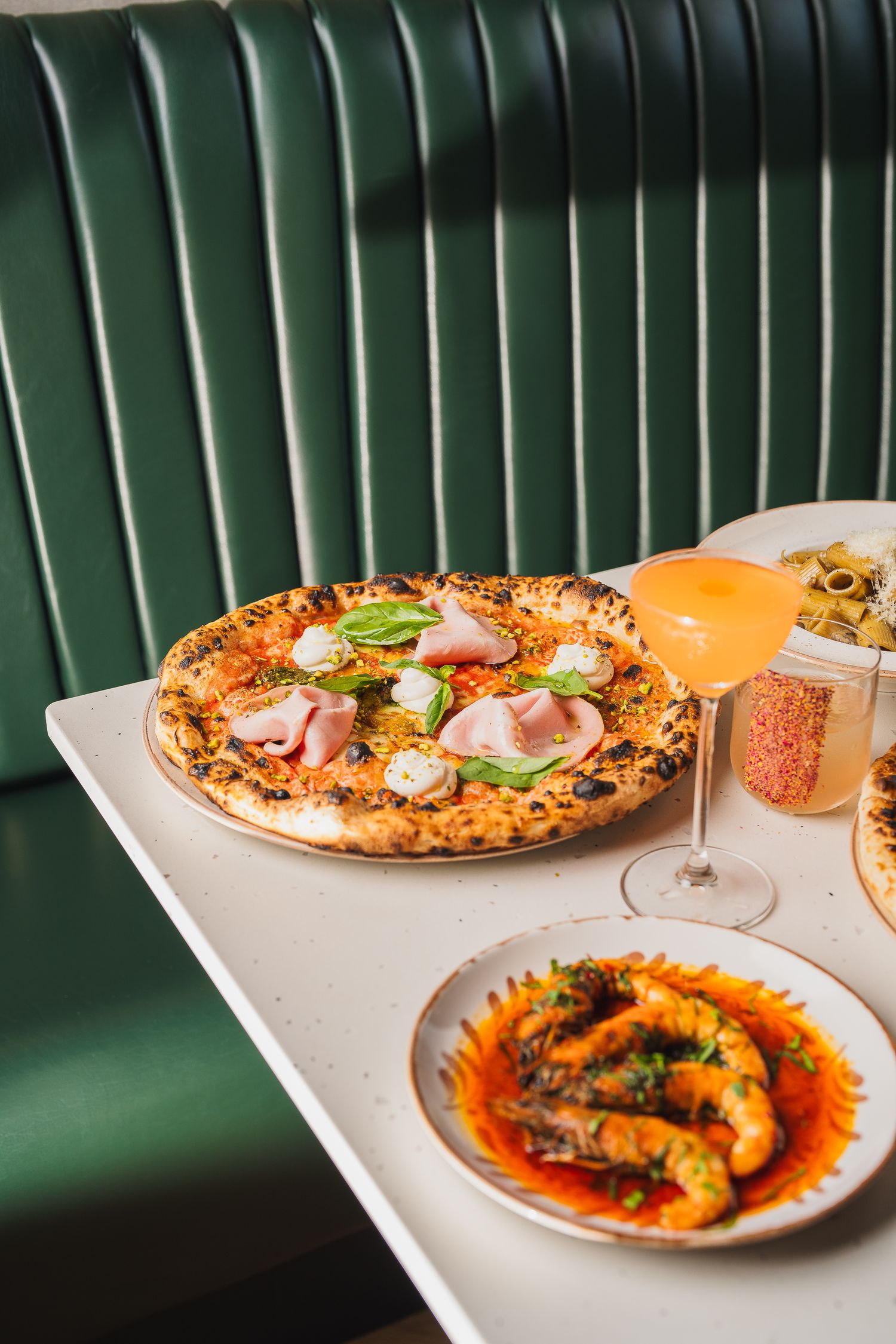Neapolitan pizza with ham and mozzarella beside an Aperol Spritz and prawns in a green booth restaurant.