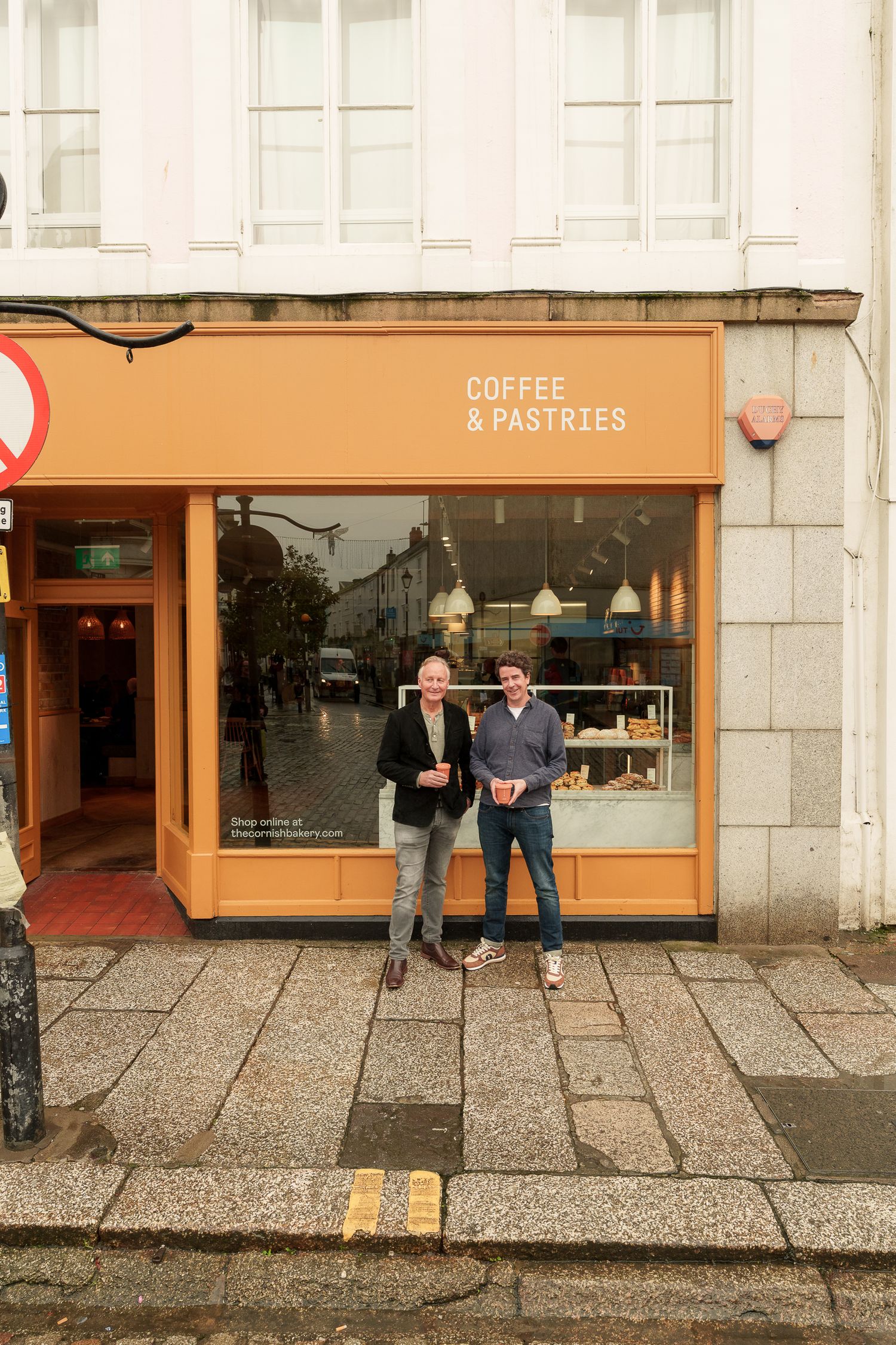 Two men standing outside an orange Coffee & Pastries cafe storefront on a rainy day.