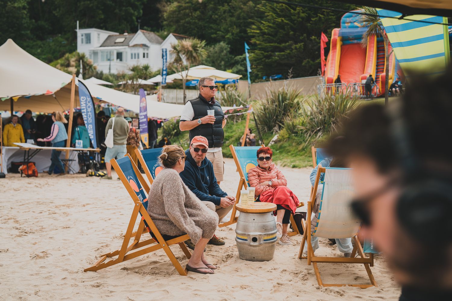 Festival goers relaxing in wooden deck chairs on sandy beach with colorful surfboards in background.