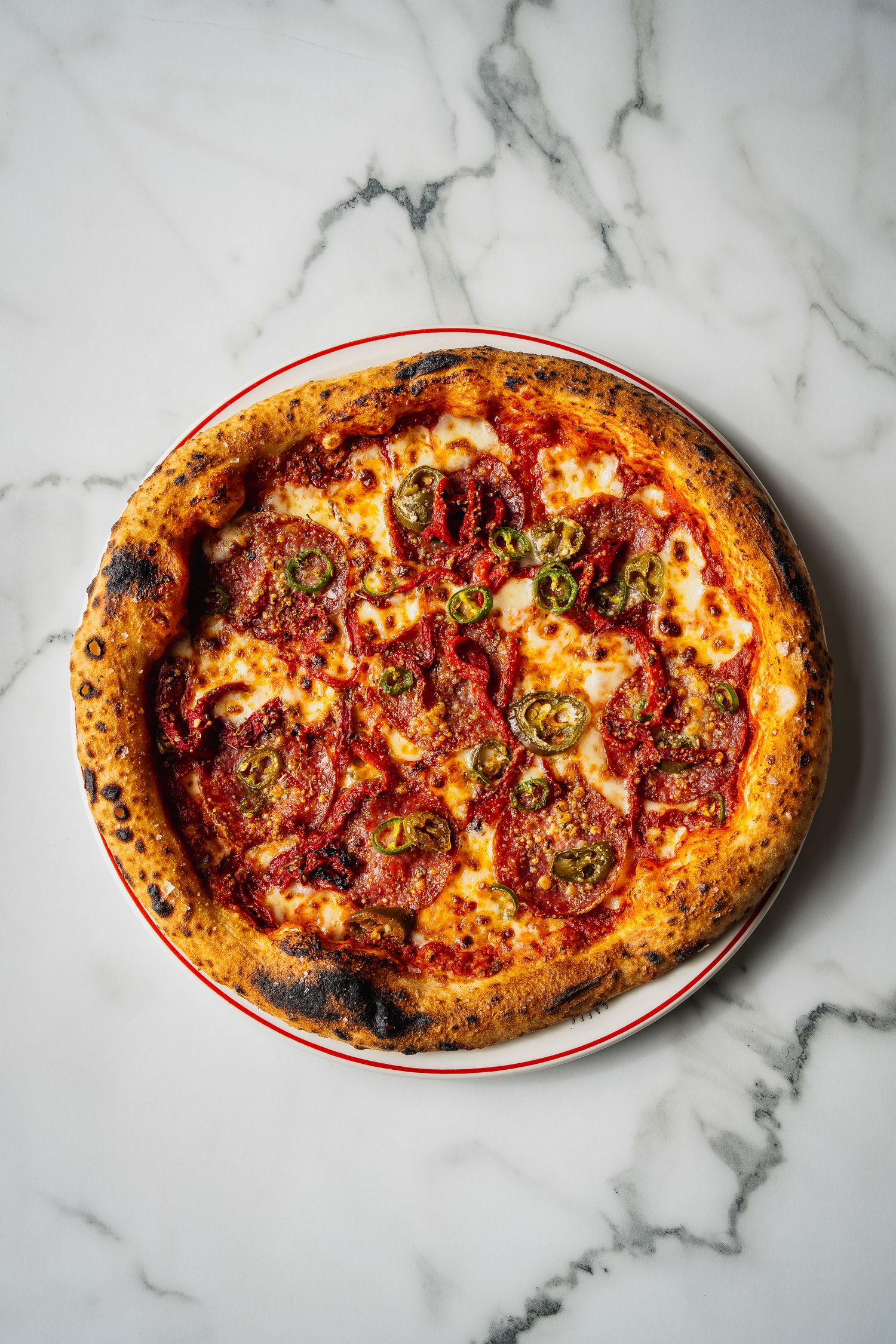 Wood-fired Neapolitan pepperoni and jalapeno pizza on marble surface with charred crispy crust.