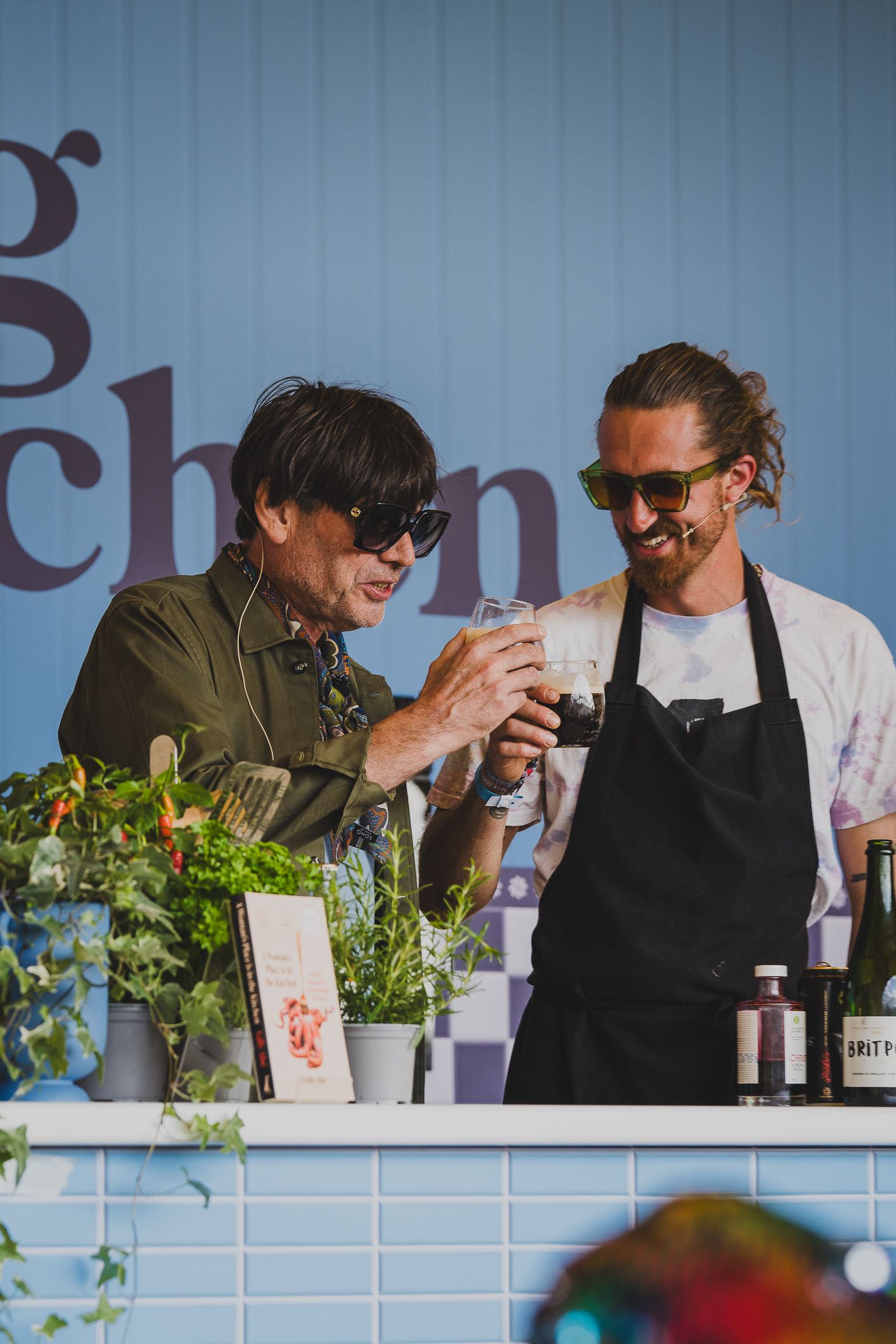 Two men wearing sunglasses toasting drinks at an outdoor kitchen bar surrounded by potted herbs.