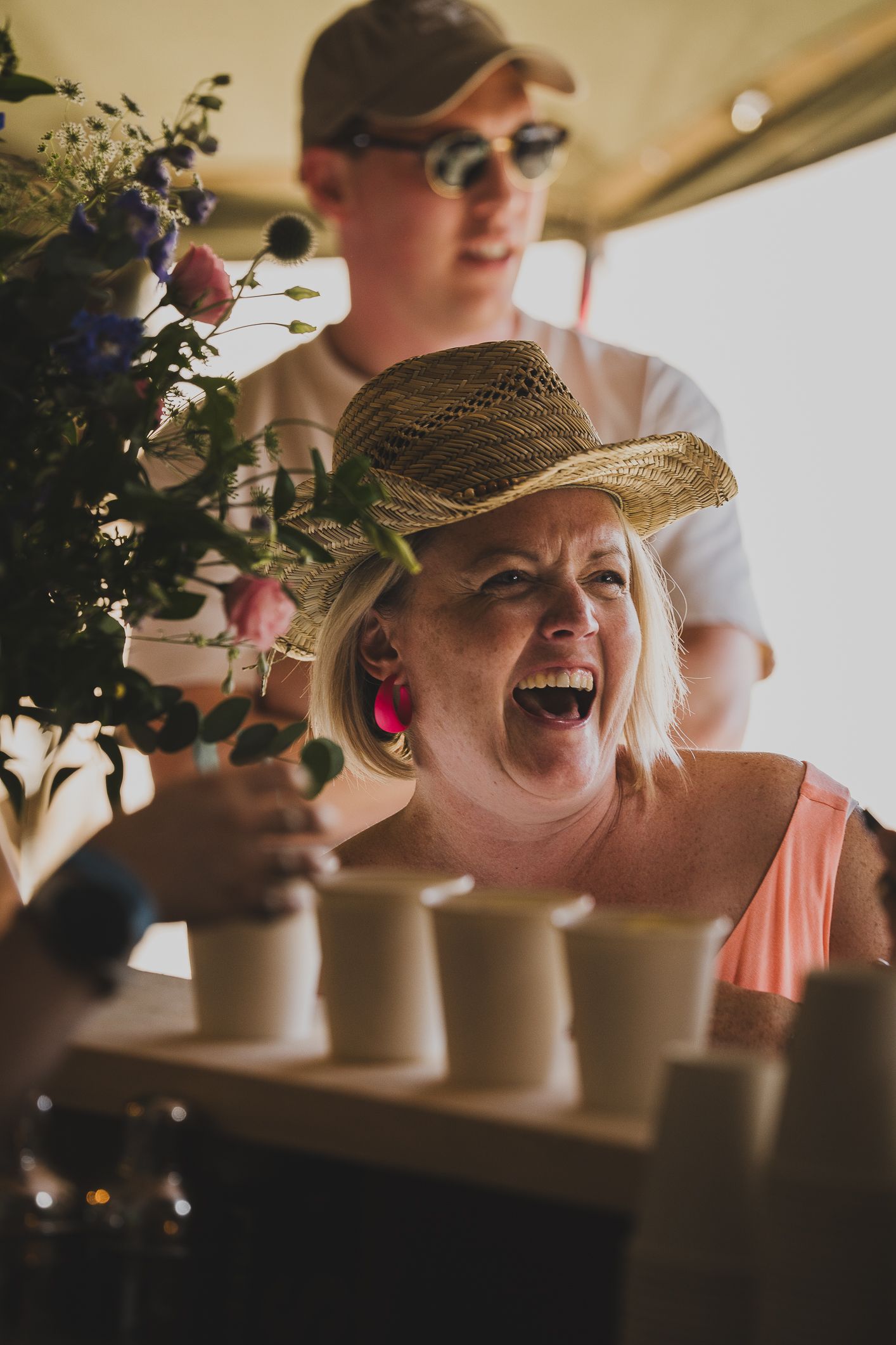 A laughing woman in a straw hat reaches for cups at an outdoor festival food stall with flowers nearby.