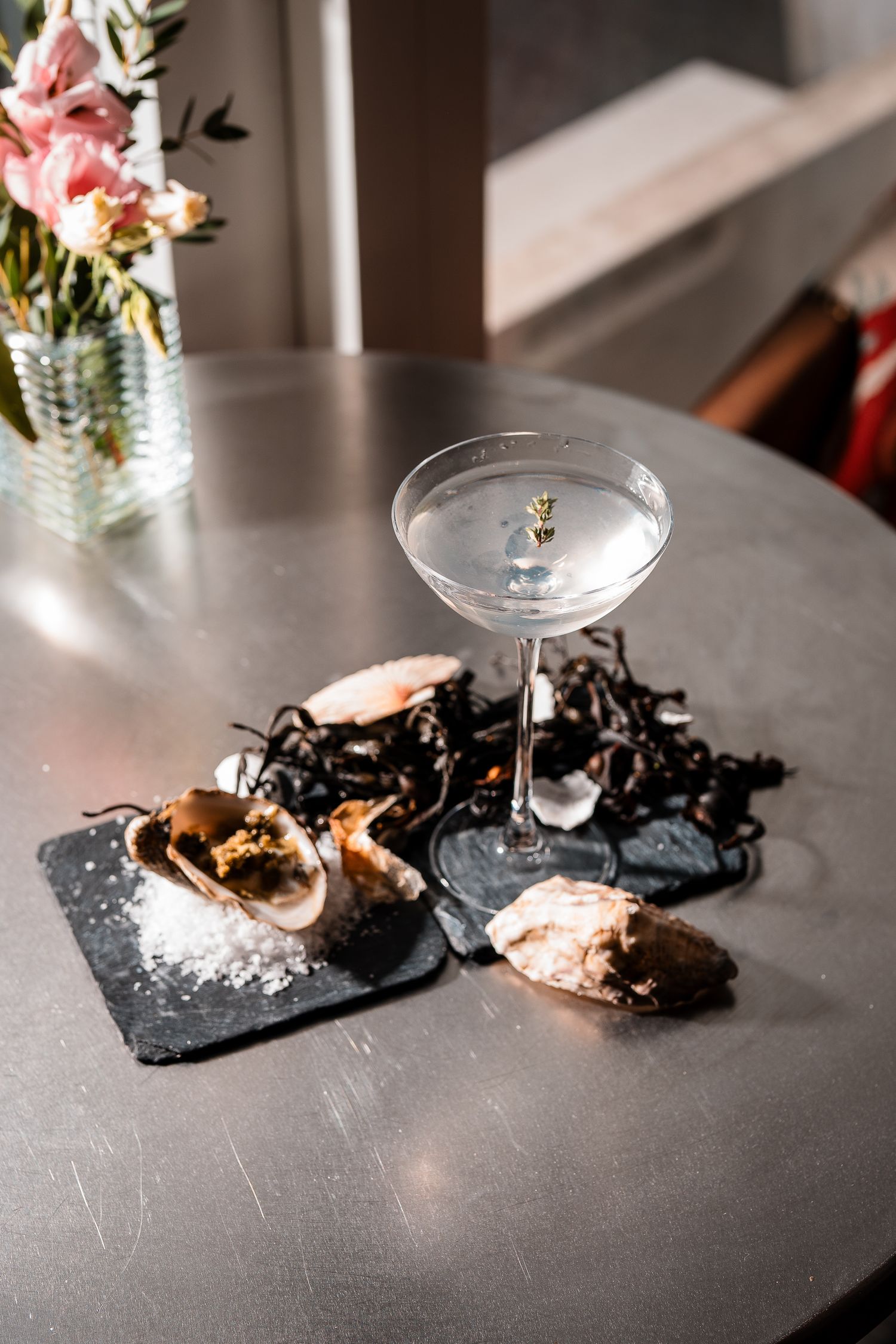 Martini glass with gold garnish surrounded by oyster shells and seaweed on slate board on round table.
