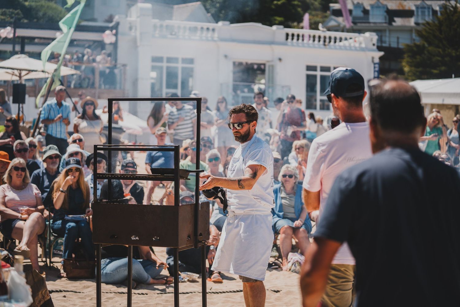 Chef in white apron cooking over open fire grill at outdoor beach food festival.