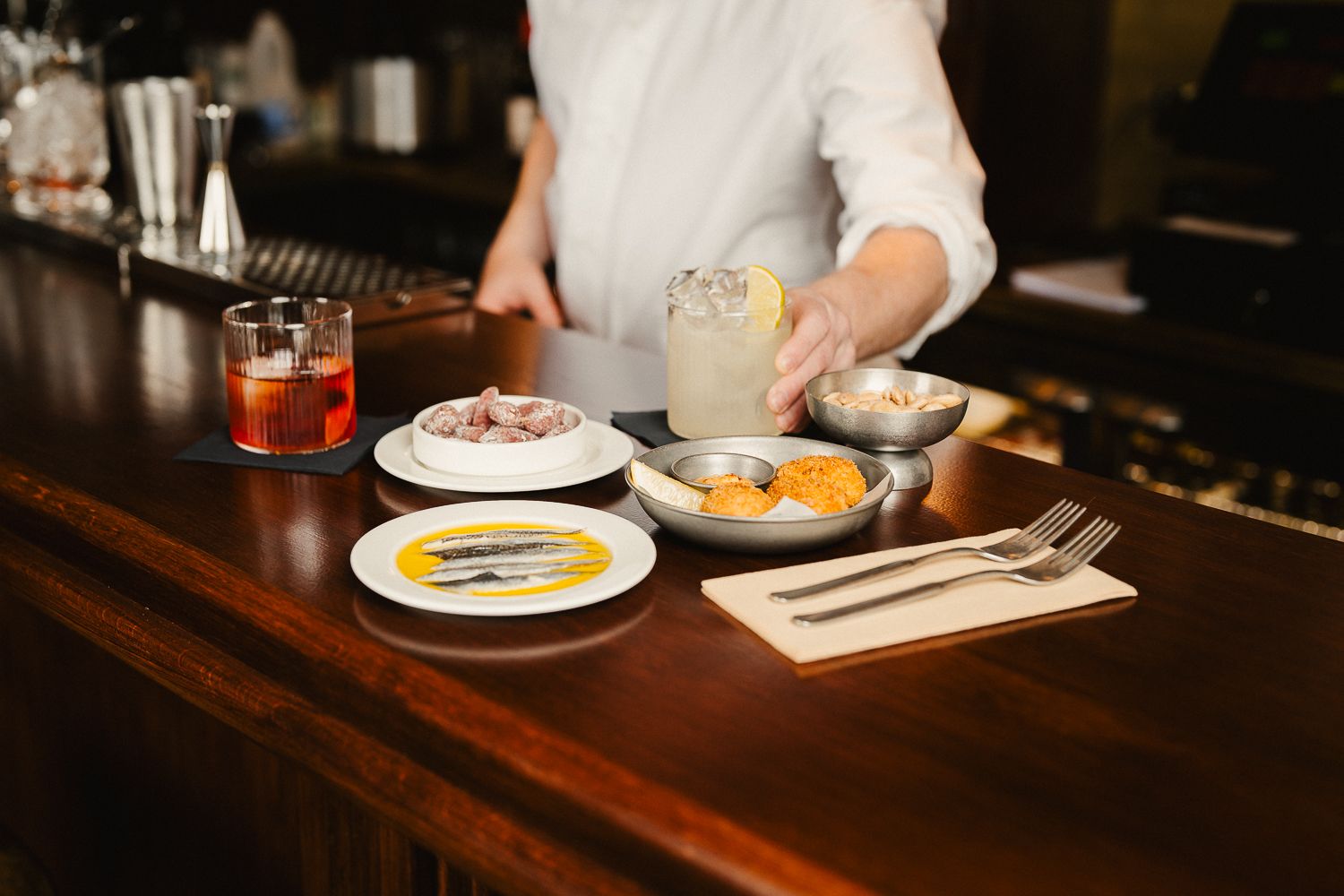 Chef presenting elegant tapas spread with anchovies, cured meats, and cocktails on a wooden bar counter.