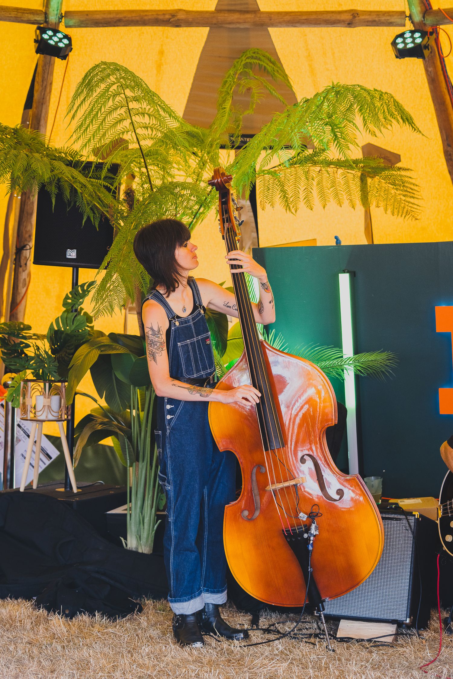 A tattooed woman in denim overalls plays a double bass on a plant-filled yellow tent stage.