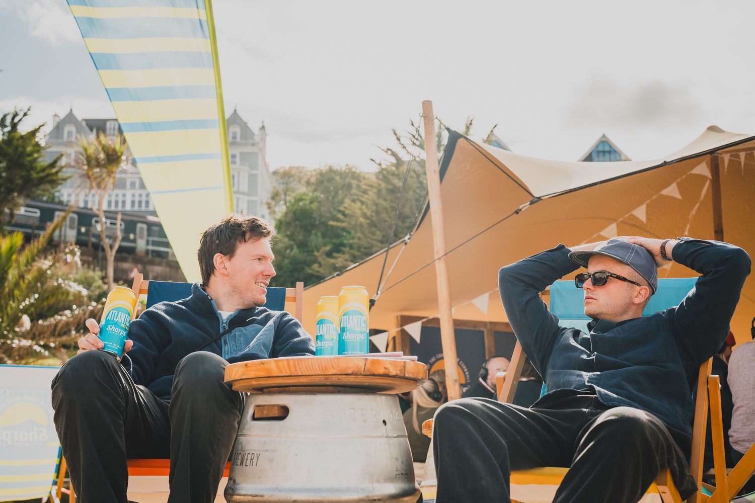 Two men relaxing in deck chairs at a sunny beach festival with colorful bunting overhead.