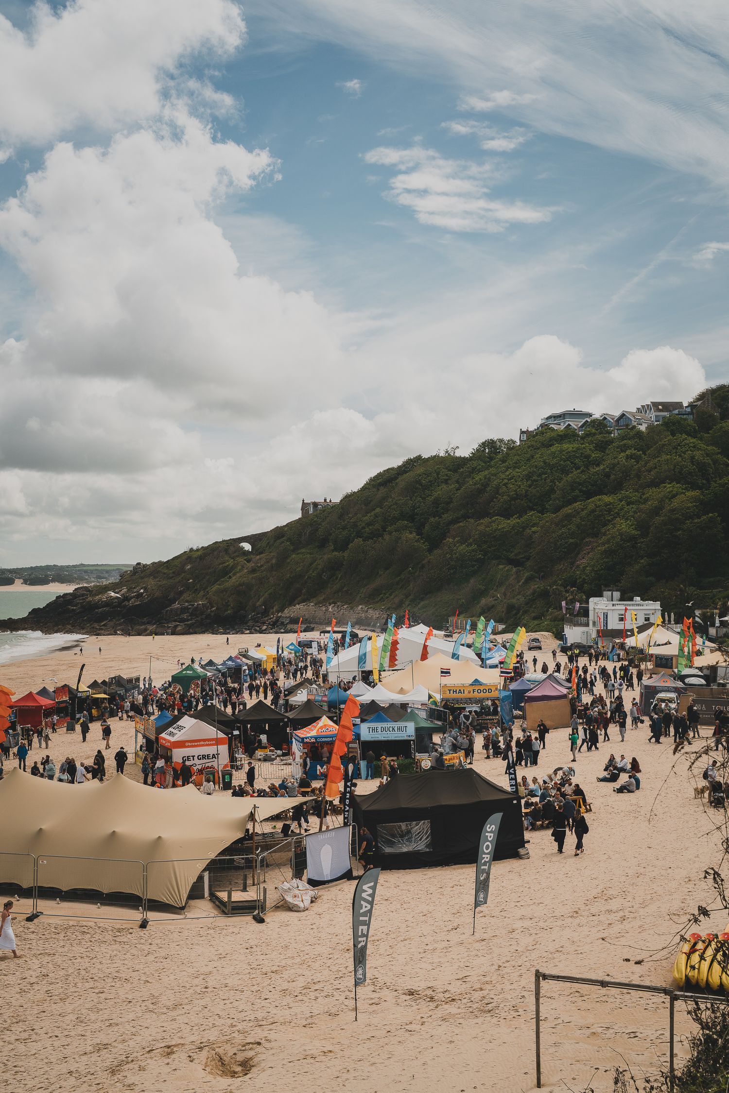Large beach festival with numerous vendor tents and flags on sandy shore beneath dramatic cloudy sky.